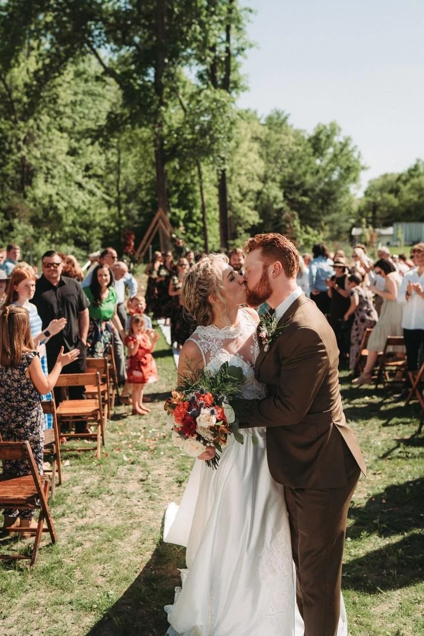 A couple kissing during their outdoor wedding ceremony surrounded by friends and family on a sunny day with green trees in the background.