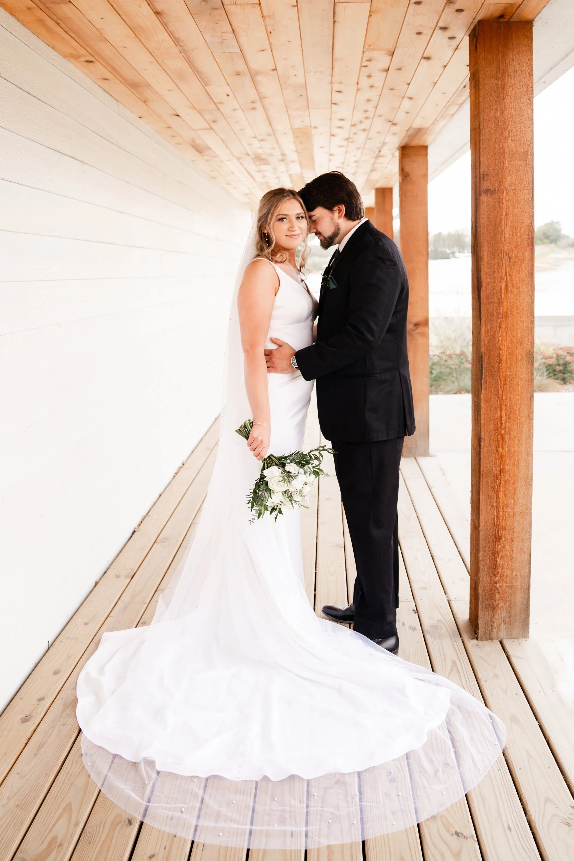 Bride and groom standing close together under a wooden canopy, with the bride holding a bouquet of white flowers and greenery, both dressed in wedding attire.