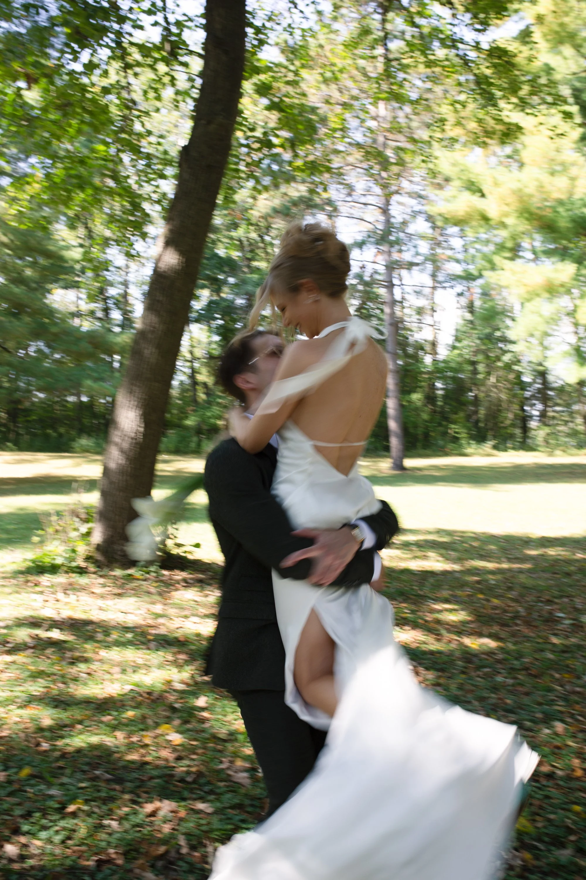 A couple, with the man in a black suit and the woman in a white wedding dress, kissing and embracing outdoors among trees, with the woman being lifted by the man, in a forest setting.
