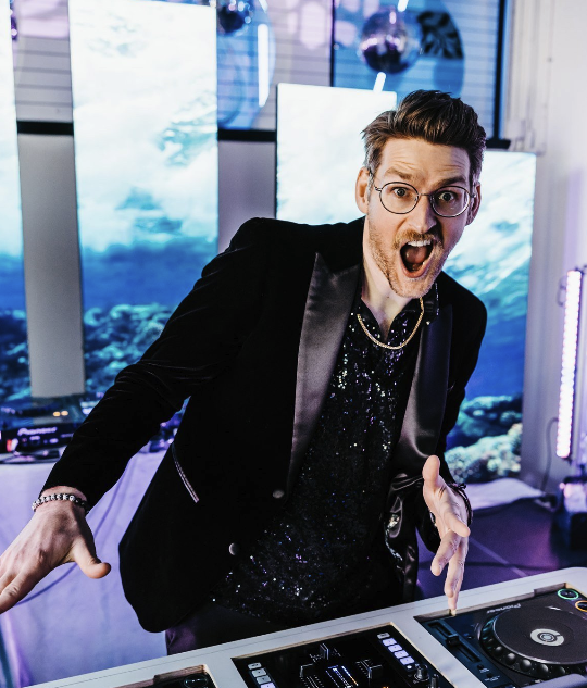 A man with glasses and a beard wearing a black tuxedo with a sequined shirt in a club or event space with aquarium tanks and screens in the background, looking excited and pointing towards the camera.
