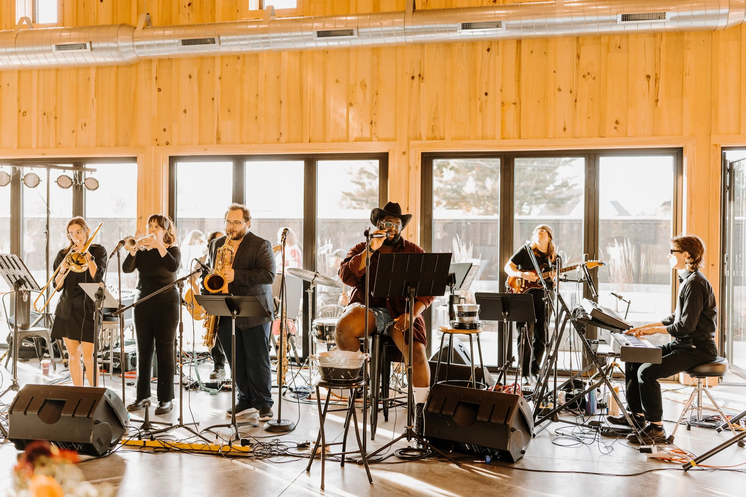A band of six musicians performing indoors with large windows in the background. The group includes a keyboardist, saxophonists, a trumpet player, a vocalist, and a guitarist. The room features wooden walls and natural sunlight.