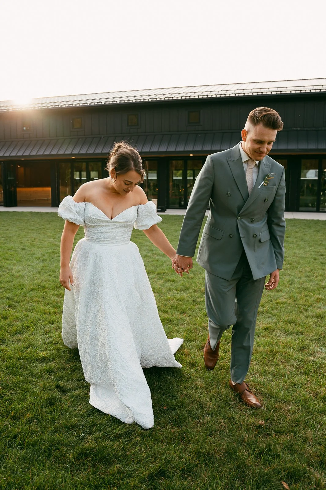 A bride and groom walking hand-in-hand on a grassy field during sunset, with a modern black building in the background.