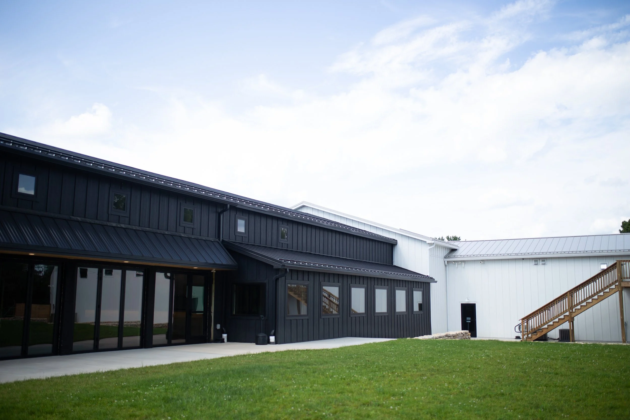 Modern building with black and white exterior walls, glass doors, and a wooden staircase outside, surrounded by a grassy lawn under a partly cloudy sky.