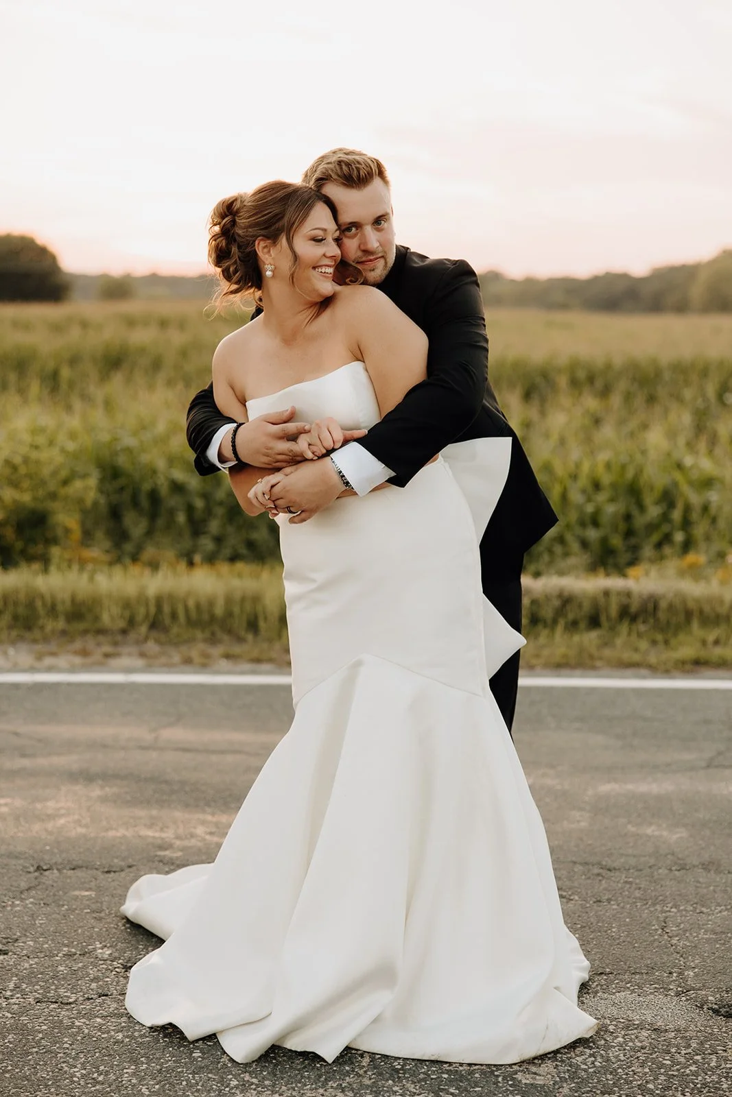 A newlywed couple hugging outdoors, the bride in a strapless white wedding gown and the groom in a black tuxedo, standing on a rural road with a grassy field and sunset sky in the background.
