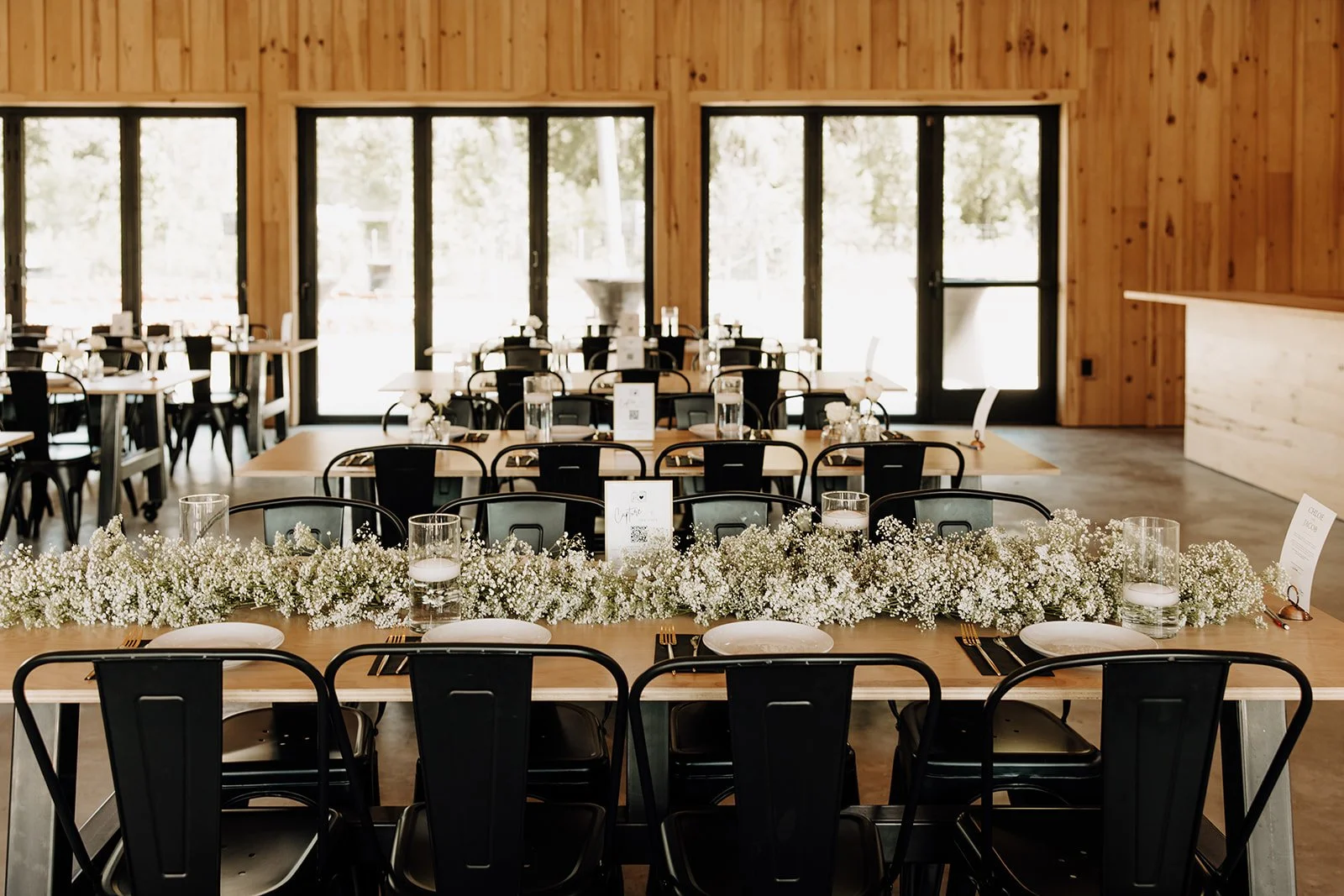 Wedding reception setup with tables decorated with white flowers, candles, and place settings, inside a wooden hall with large windows.