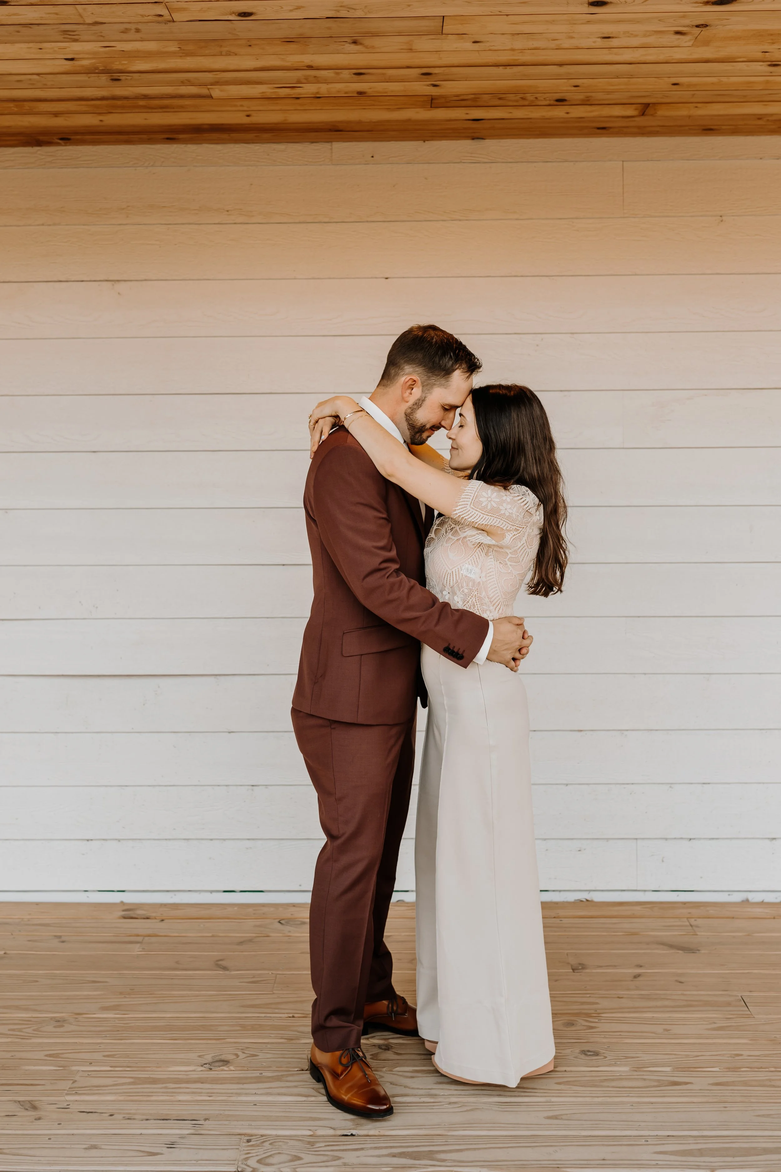 A couple is embracing each other in a loving manner, standing face-to-face with their foreheads touching against a backdrop of white wooden wall and wooden ceiling.