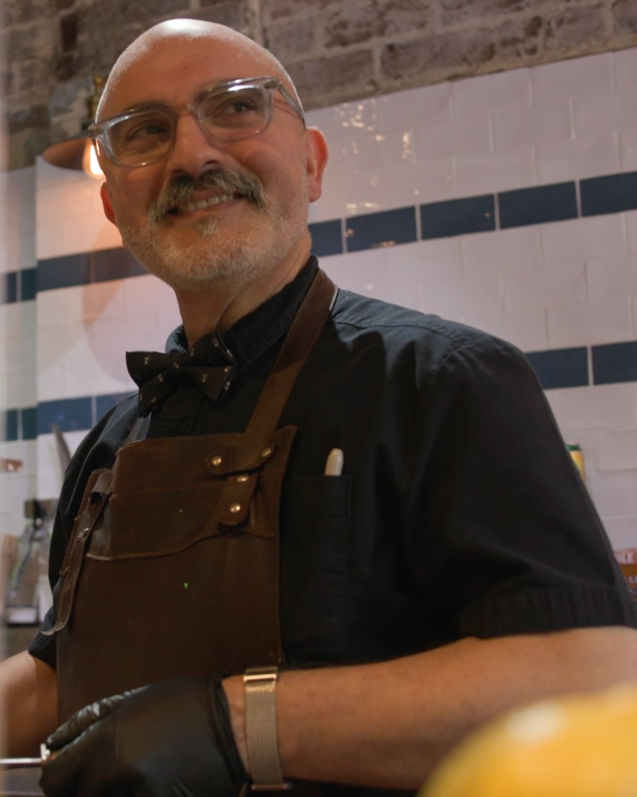 Smiling man with glasses and beard wearing a black shirt, bow tie, and brown apron, in a kitchen with white and dark blue tile walls.