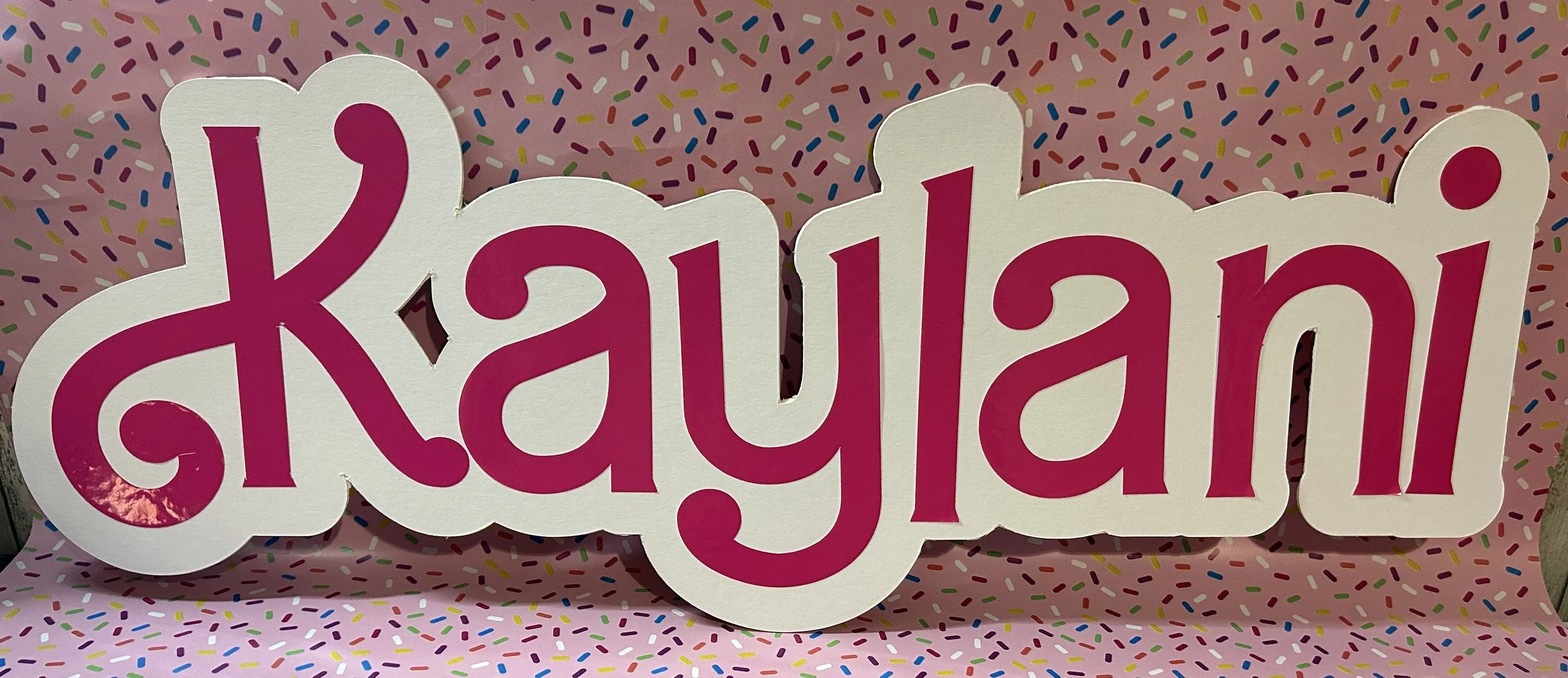 Pink paper cutout with the name "Kaylani" on a colorful sprinkle-patterned background.