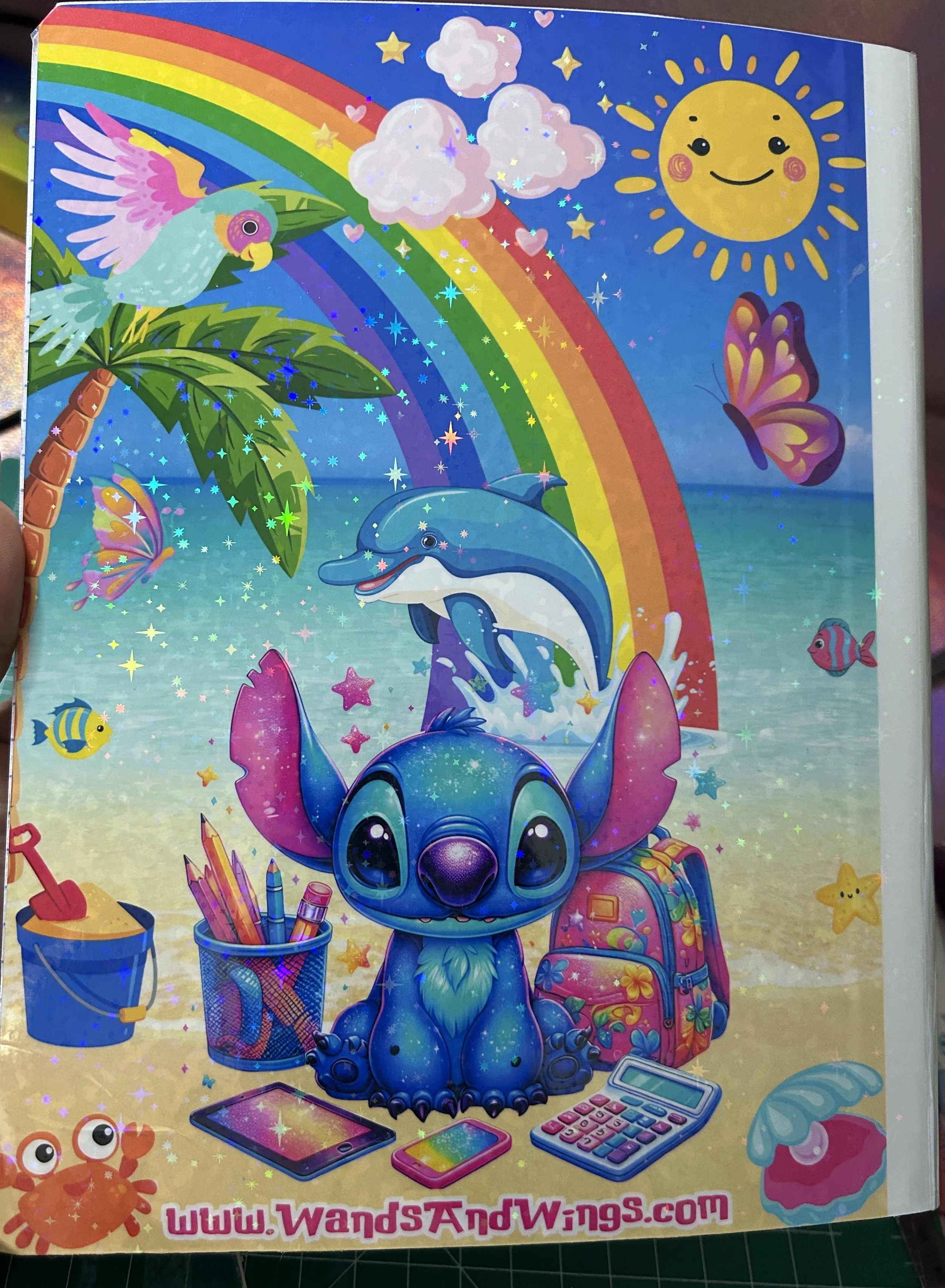 Colorful beach scene with a cute blue cartoon character, dolphin, rainbow, palm tree, smiling sun, and beach items like pencils, a backpack, calculator, and crab. Website URL "www.WandsAndWings.com" is shown at the bottom.