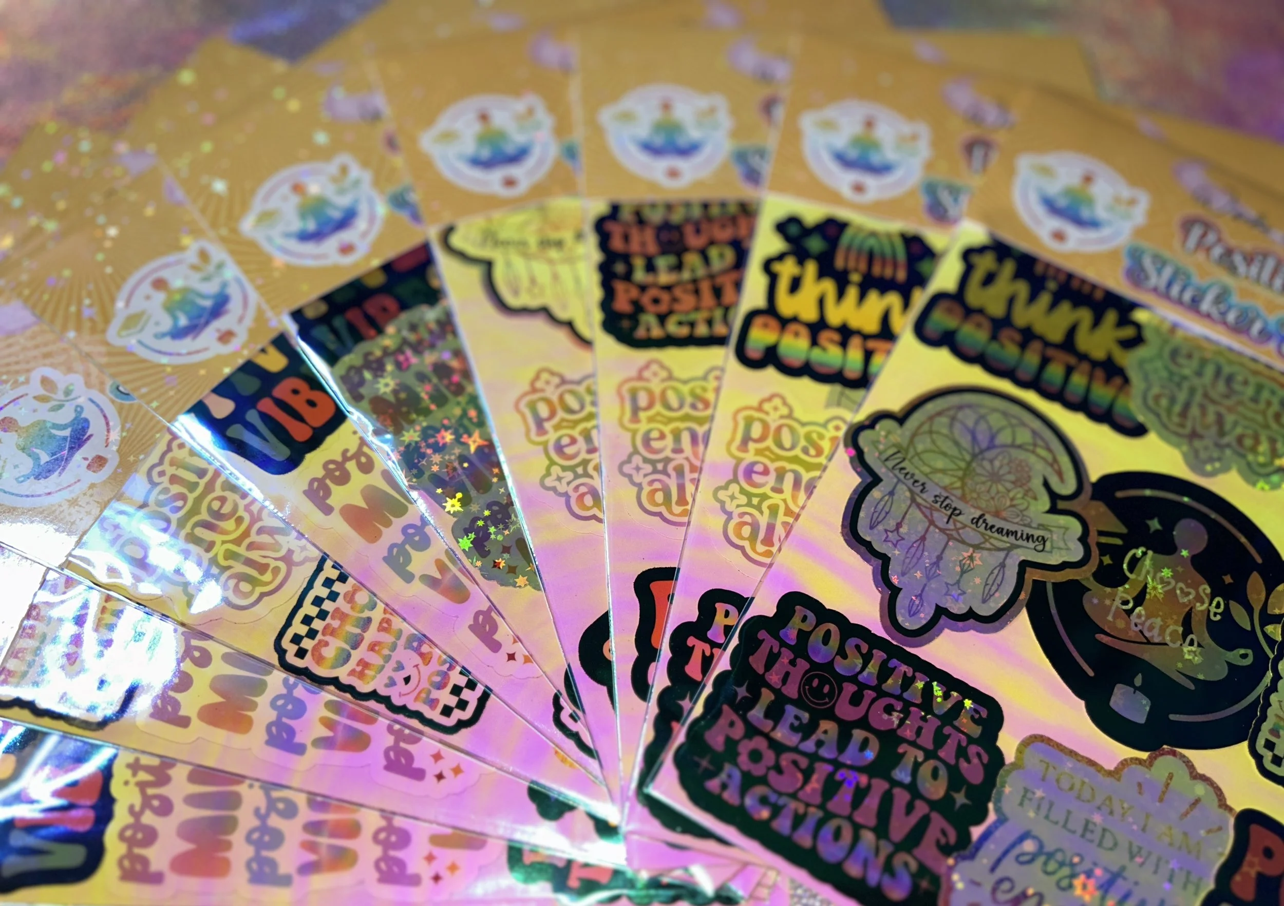 A fan arrangement of colorful sheets of holographic stickers with motivational messages such as "positive energy always" and "never stop dreaming."