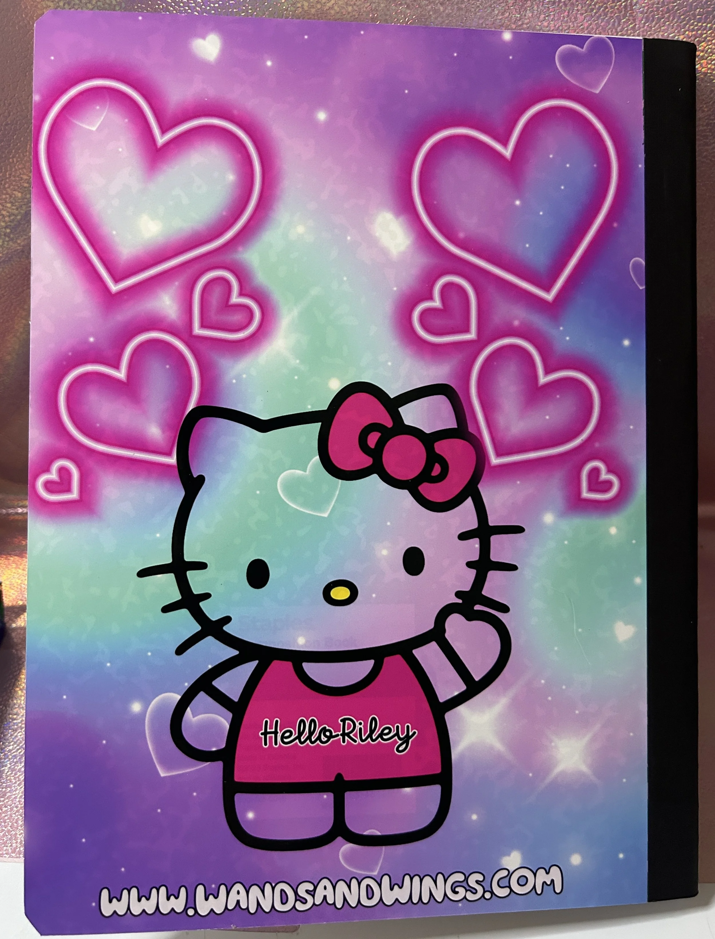 Colorful notebook cover featuring a cute cat character with a bow, surrounded by pink and purple hearts. The words "Hello Riley" are on its shirt, with the website link "www.wandsandwings.com" at the bottom.