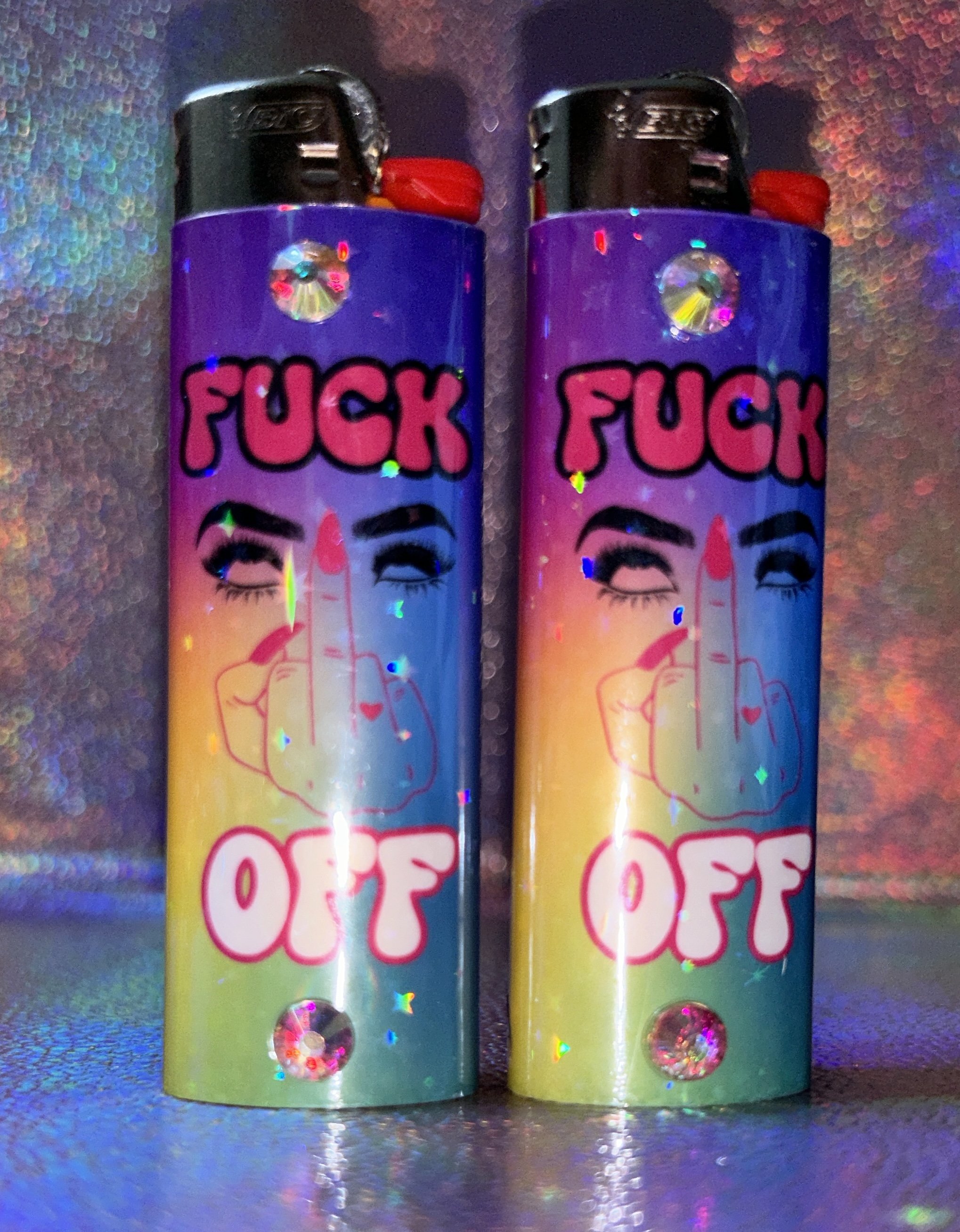 Two colorful lighters with a graphic showing a hand flipping the middle finger and text saying "FUCK OFF" against a gradient background, adorned with small decorative gems.