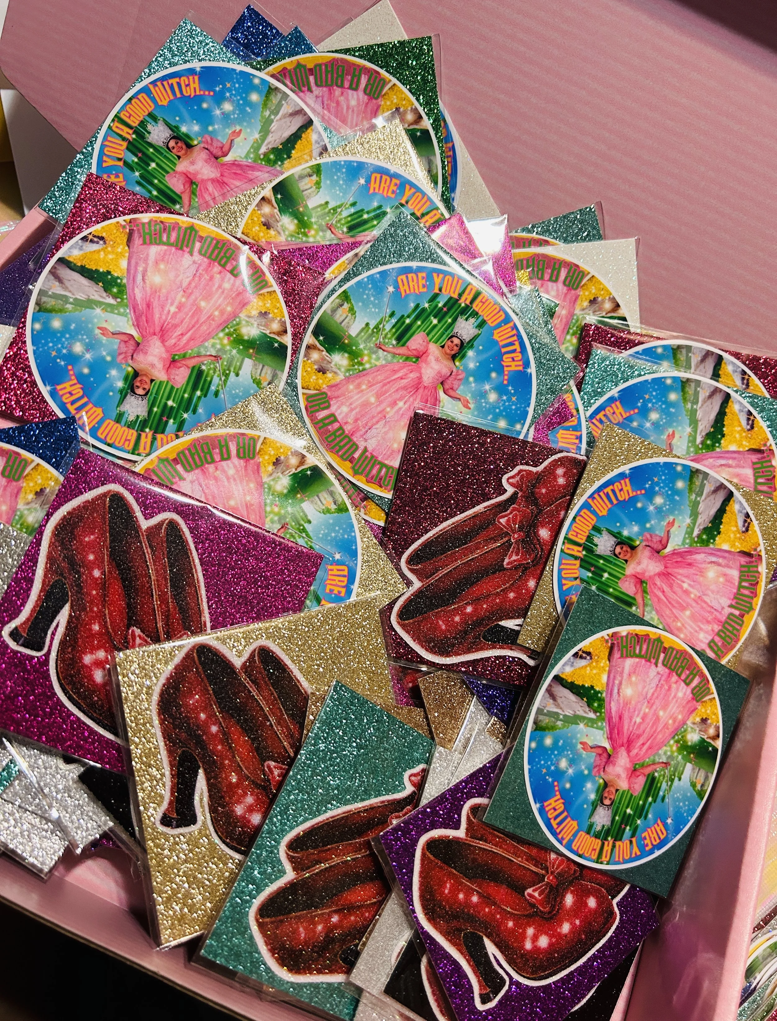 Box filled with glittery stickers featuring images of ruby slippers and the phrase 'Are You A Good Witch?' with a pink dress and background.