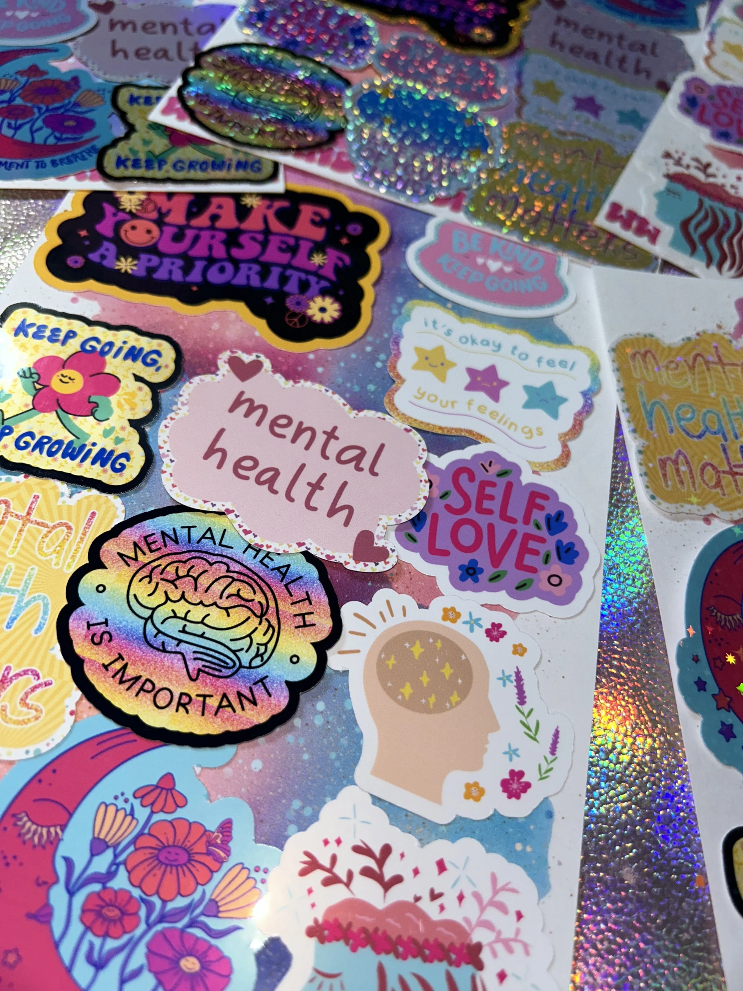 Collection of colorful mental health stickers with positive messages like "Make Yourself a Priority," "Self Love," and "Mental Health is Important." Background has a holographic pattern.
