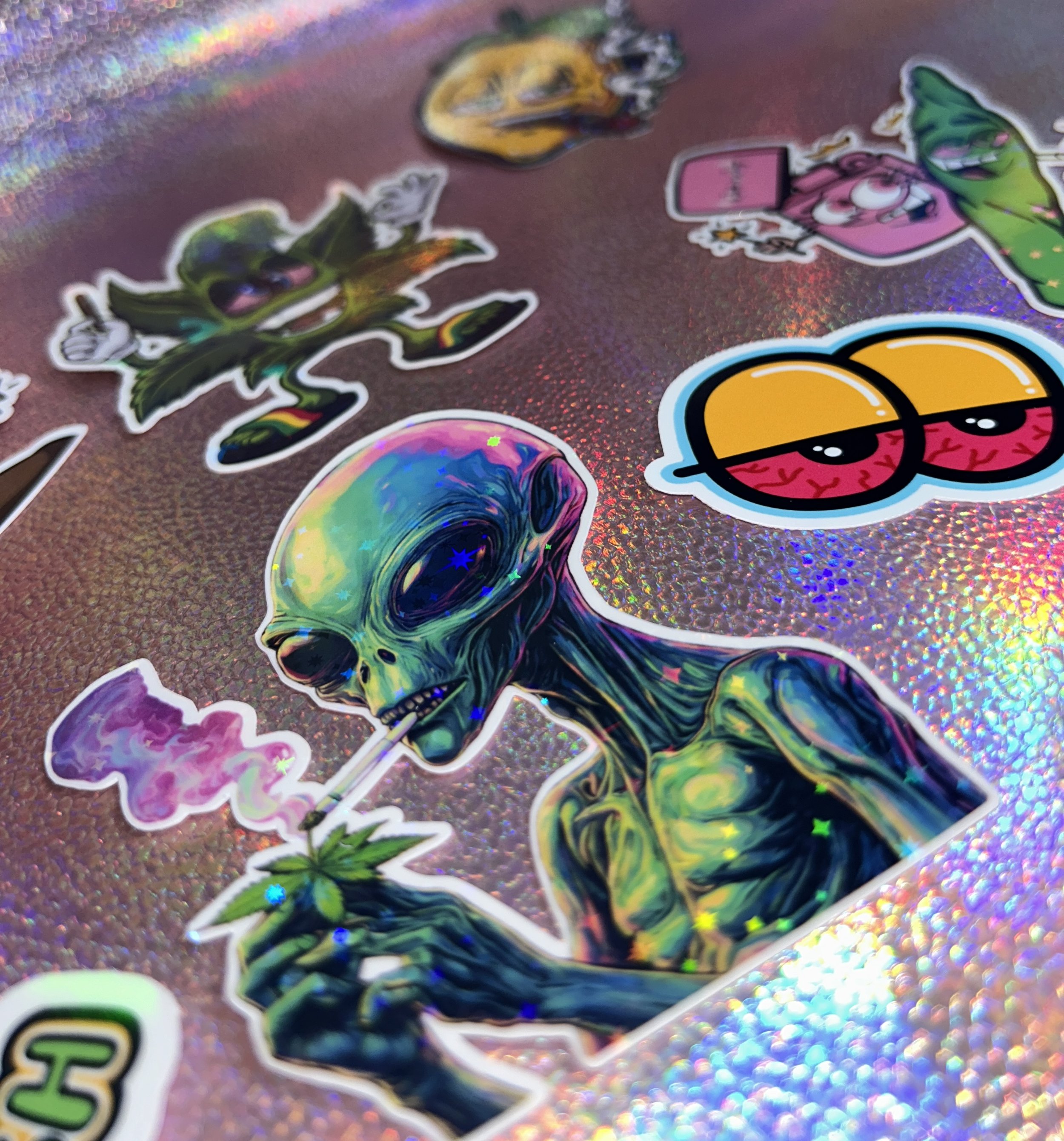 Colorful stickers featuring an alien smoking, a character with leaves, and cartoonish eyes on holographic background.