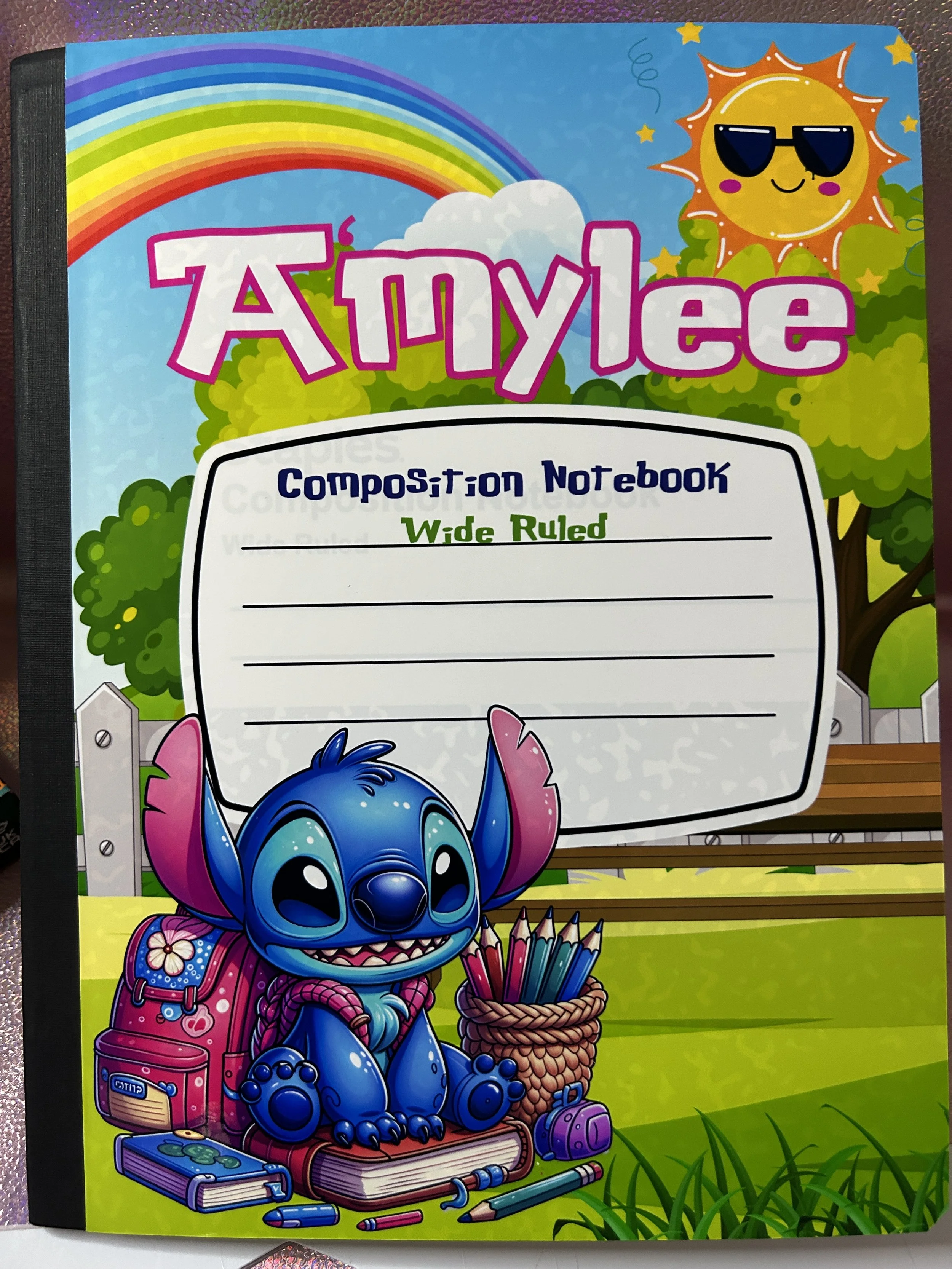 Colorful composition notebook cover featuring a cartoon character sitting on books, holding school supplies. A rainbow arcs across the top with a sun wearing sunglasses, and trees in the background. The word "Amylee" is prominently displayed at the t