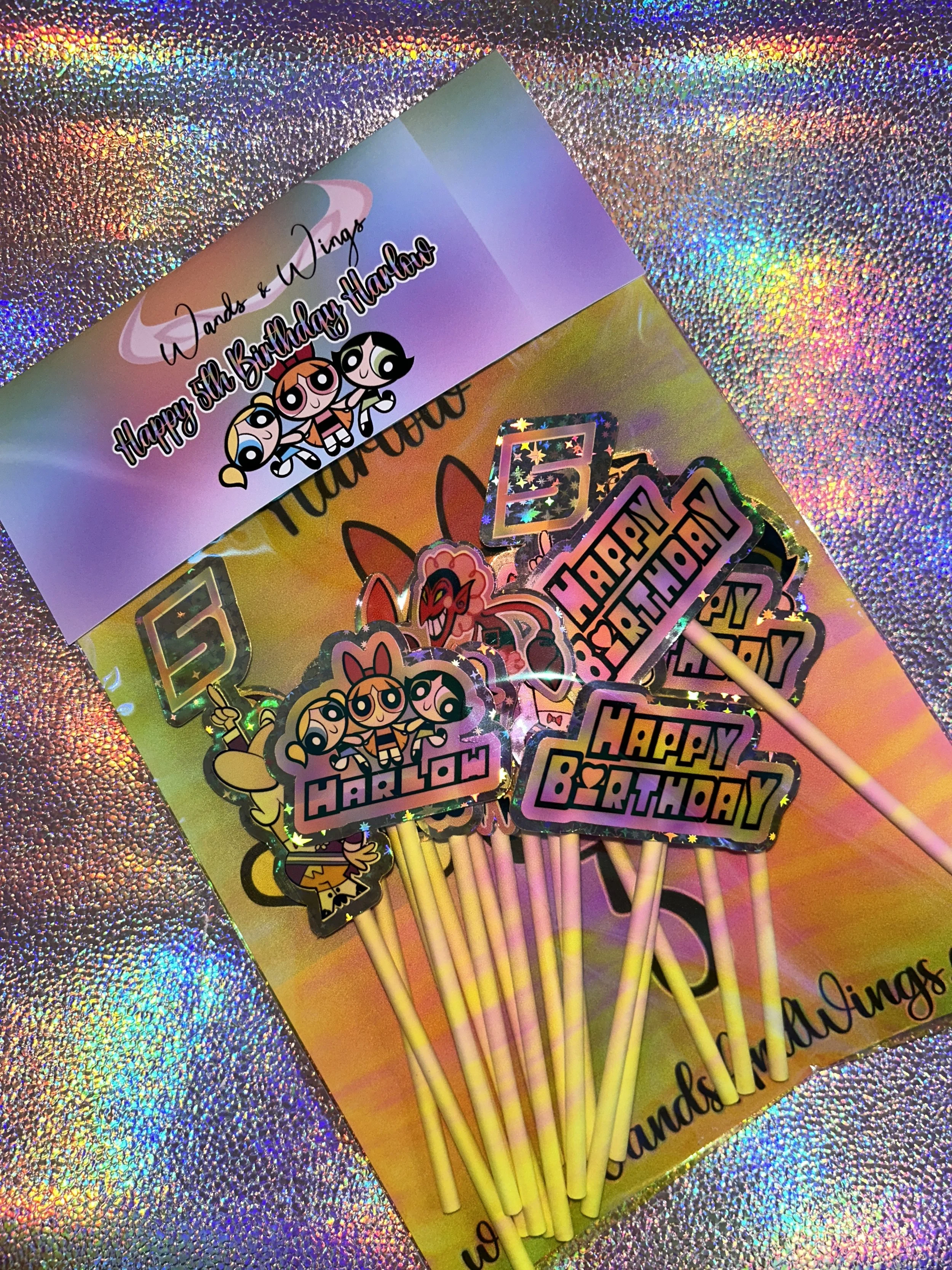 Birthday party decorations featuring Powerpuff Girls themed cupcake toppers on a holographic background. Includes "Happy Birthday" and "Harlow" designs with number 5 cutouts.