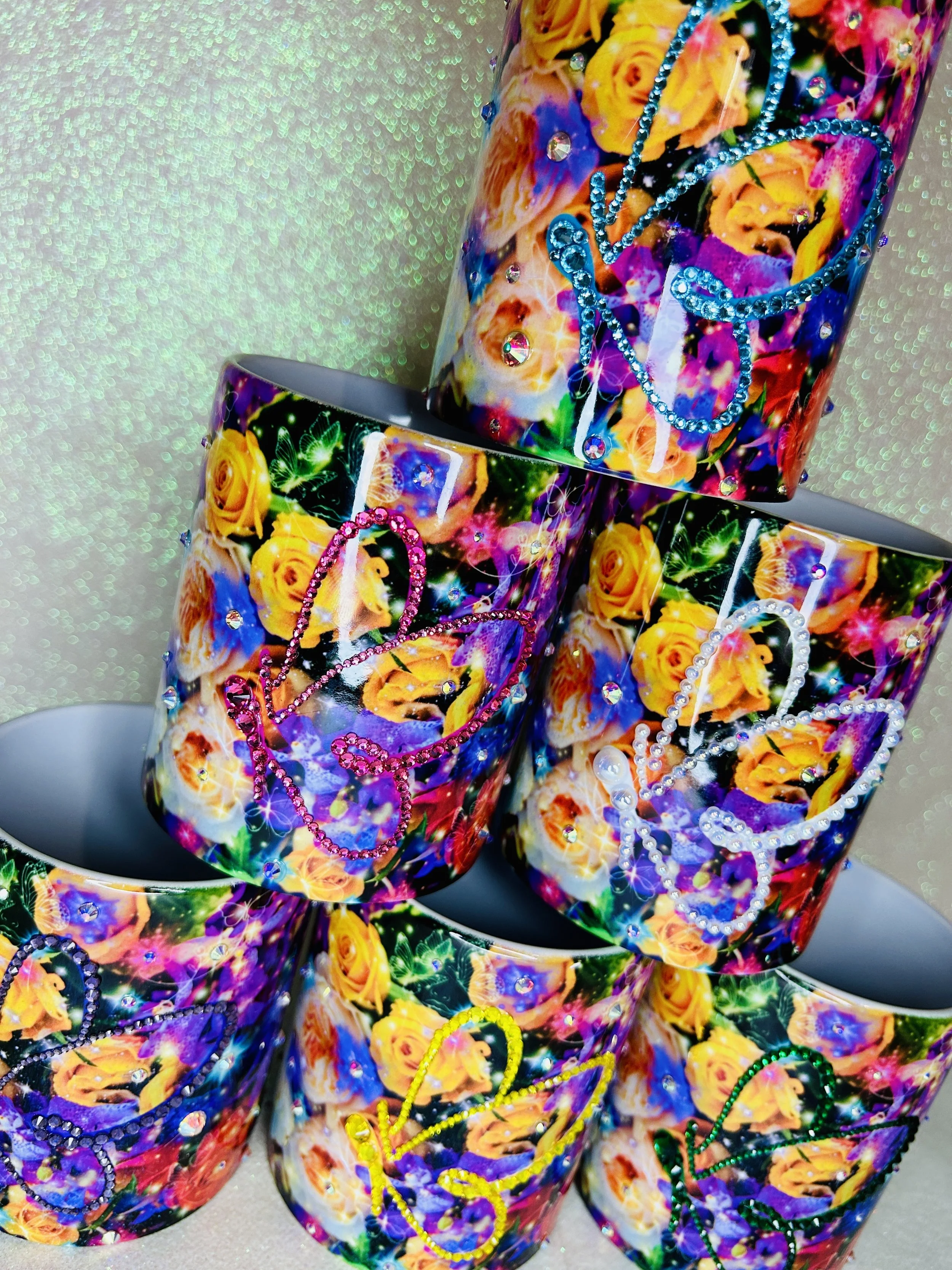 Colorful mugs with floral patterns and rhinestone heart designs, stacked against a glittery background.