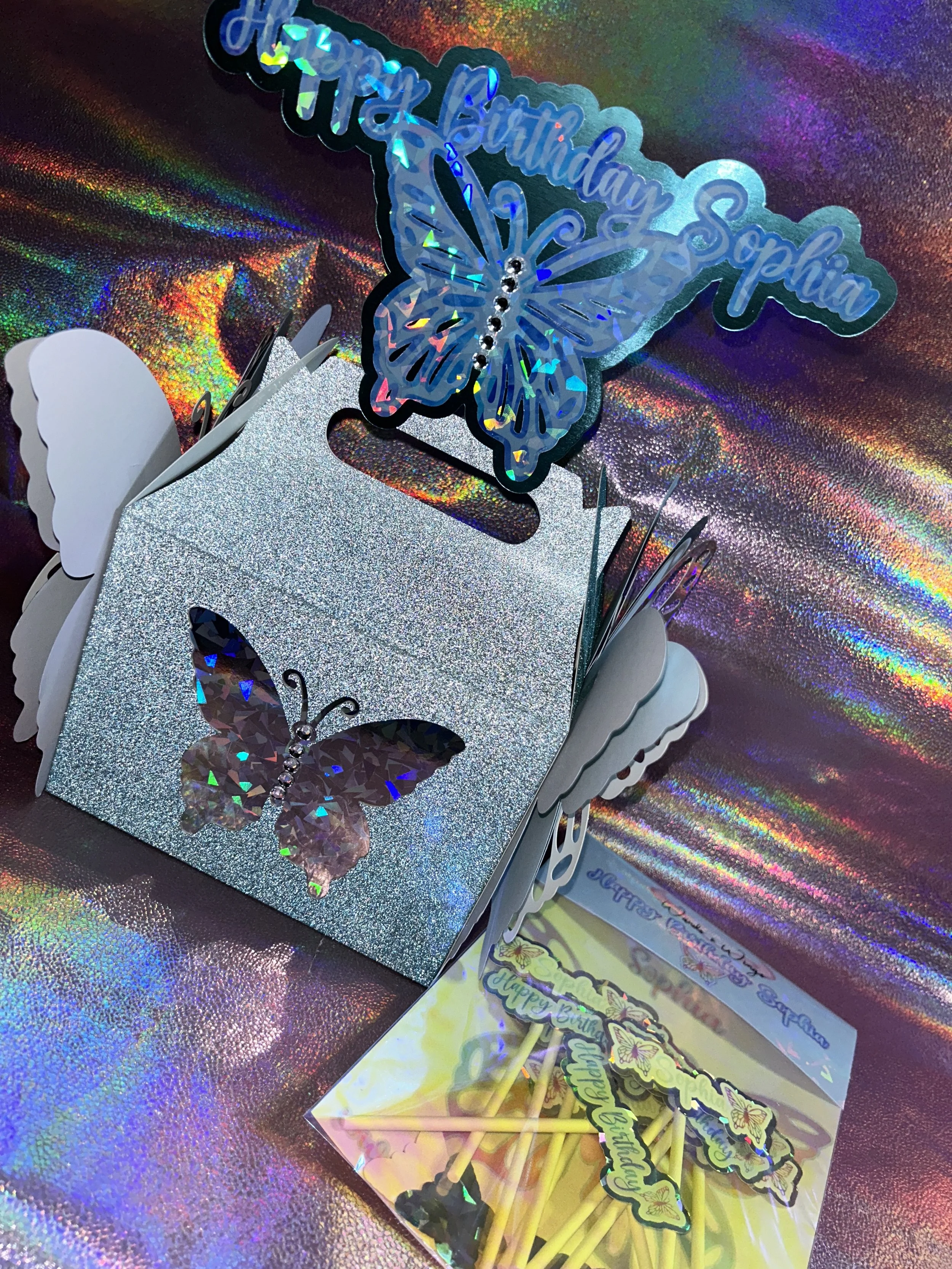 Holographic birthday decoration with butterfly theme, including a glittery gift box and cupcake toppers.