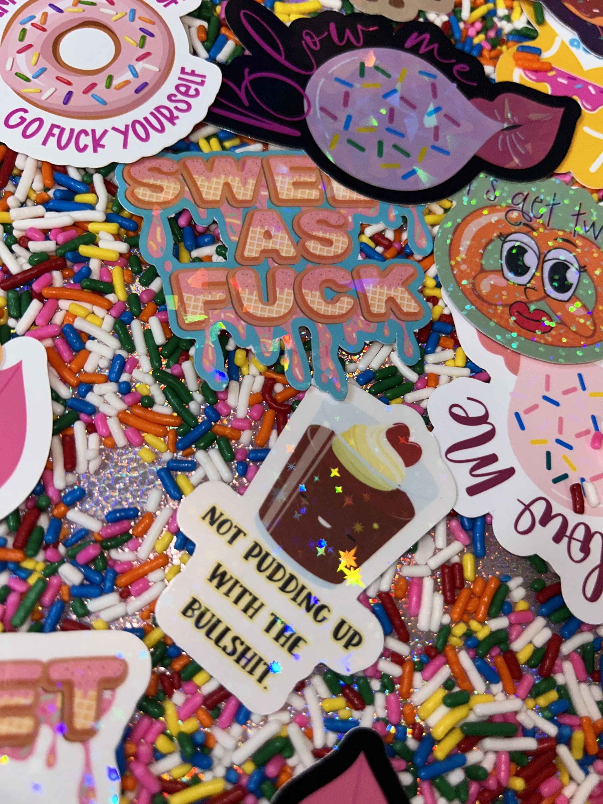 A collection of colorful stickers featuring playful, irreverent messages and dessert-themed designs, set against a background of rainbow sprinkles. Notable stickers include phrases like "Sweet As F***" and "Not Pudding Up With The Bullsh**."