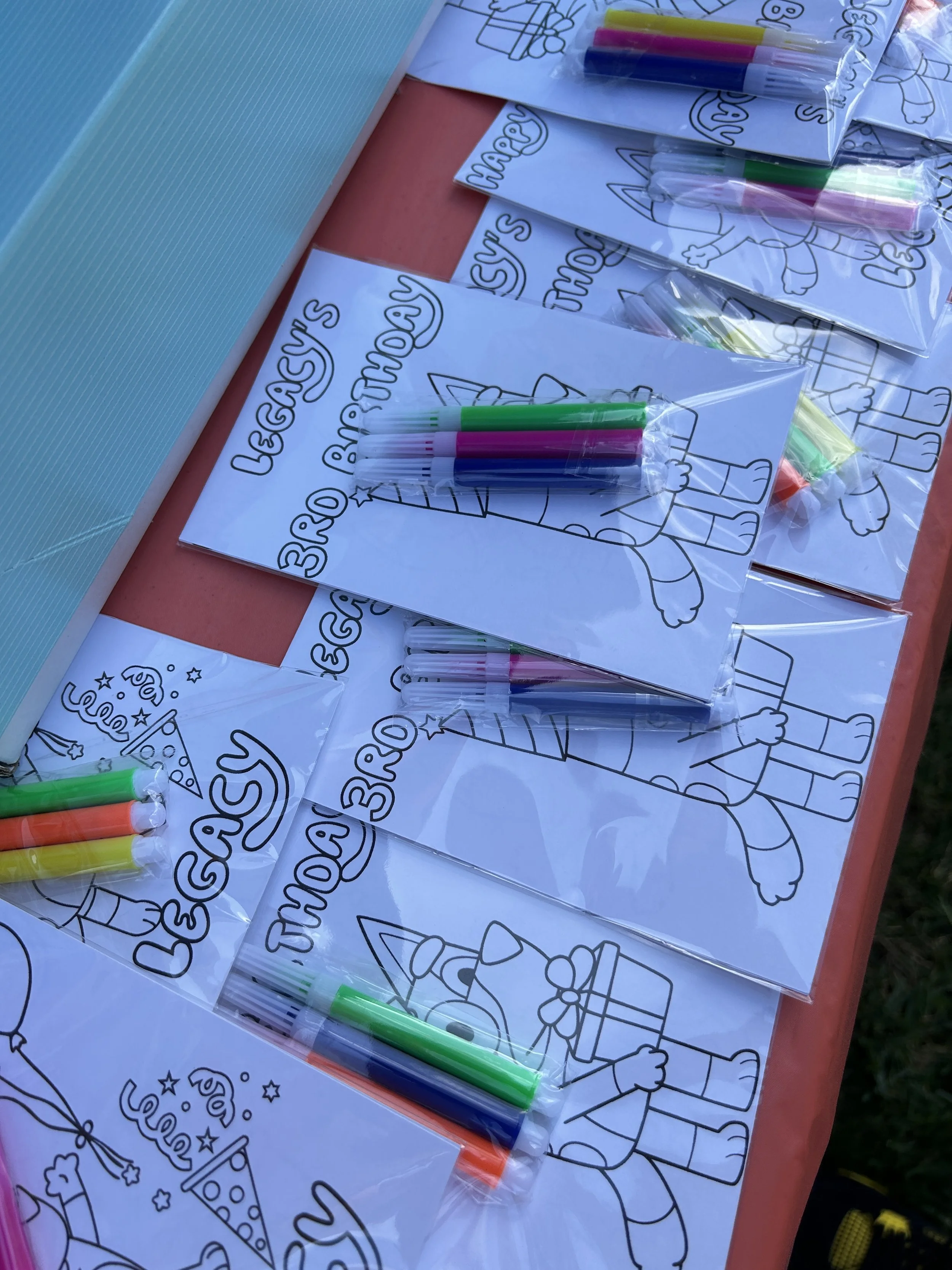 Children's coloring sheets with party drawings for a 3rd birthday, alongside small packs of colorful markers on a table.