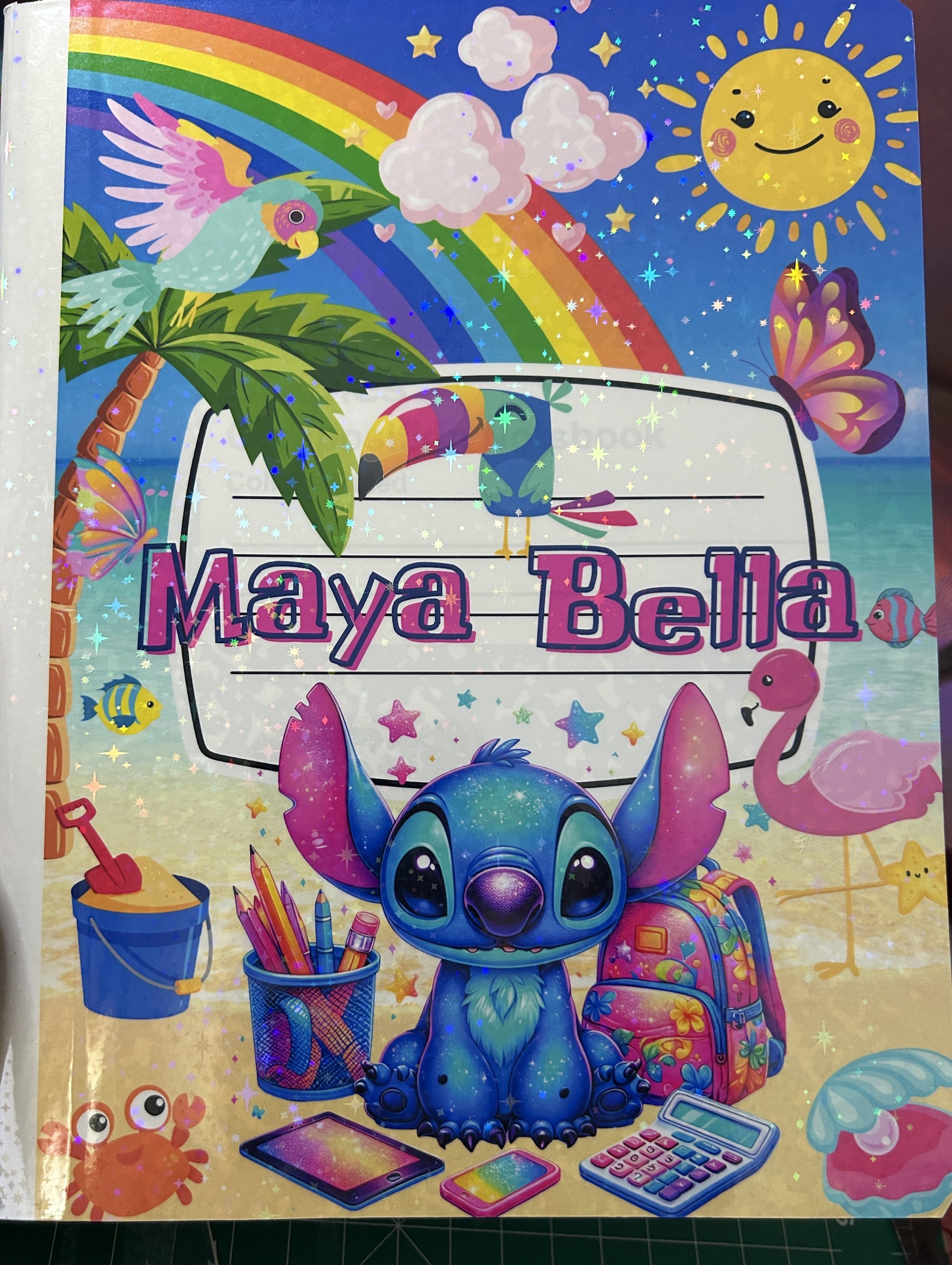 Colorful notebook cover featuring a blue alien character surrounded by school supplies, rainbow, sun, clouds, butterfly, parrot, flamingo, and beach scene with bucket and crab.