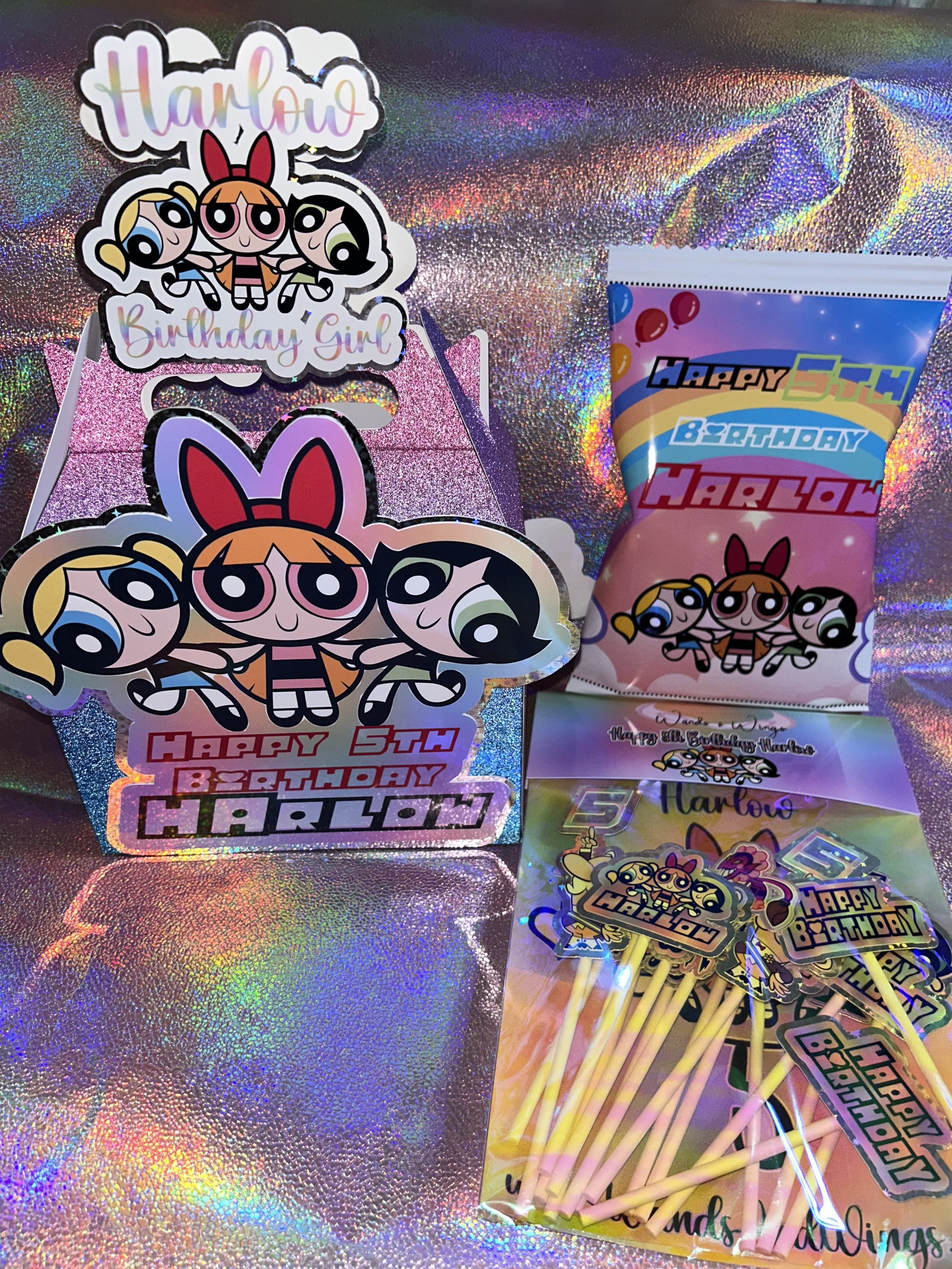 Powerpuff Girls themed birthday decorations for Harlow's 5th birthday. Includes a party favor box and cupcake toppers with character images and text "Happy 5th Birthday Harlow." Background features a colorful, iridescent surface.