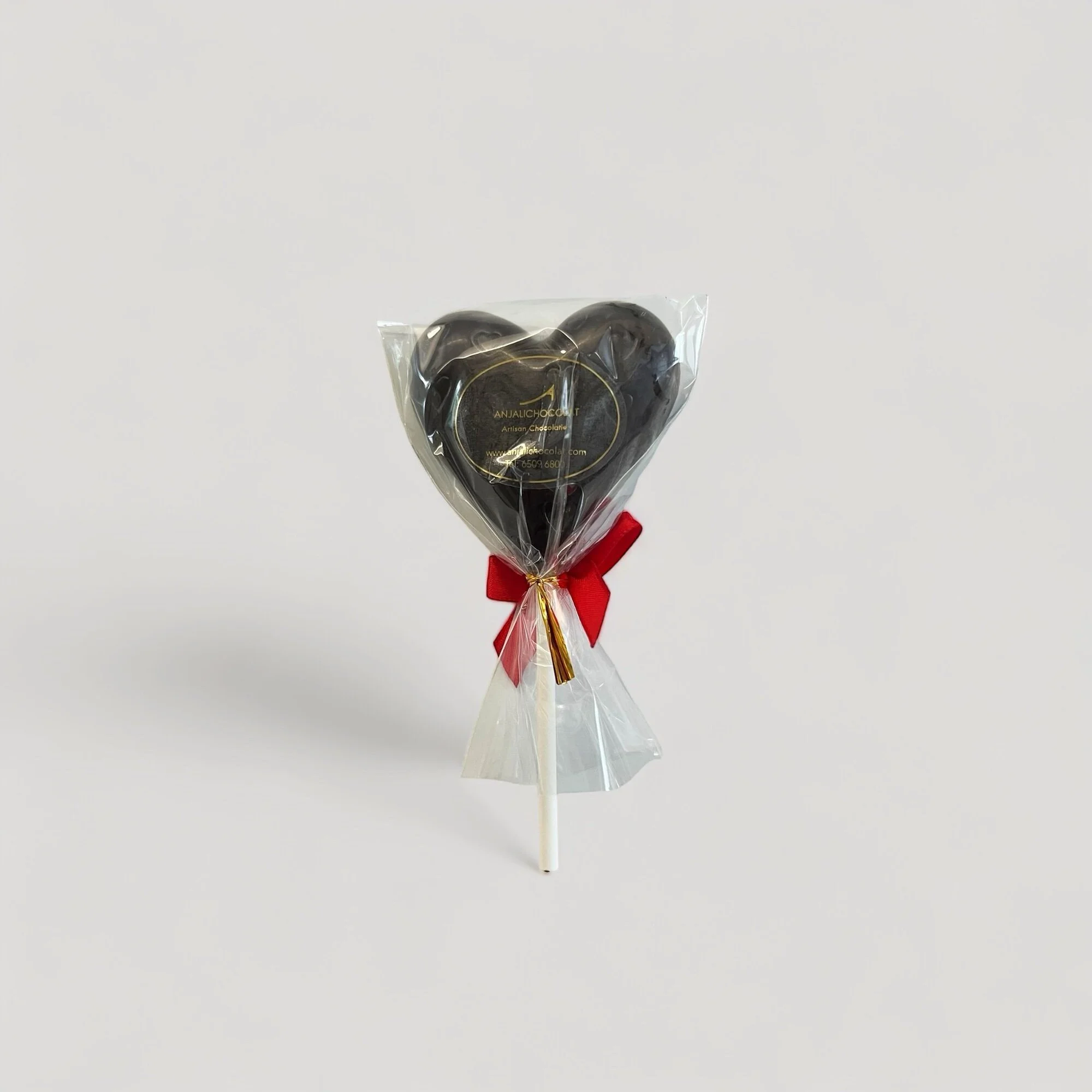 One heart-shaped lollipop in dark chocolate with a red ribbon on a light grey background. A sweet treat for Valentine's Day or any occasion