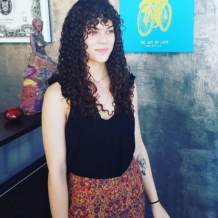 A young woman with curly dark hair wearing a black sleeveless top and a patterned skirt standing indoors against a gray textured wall with artwork and sculptures in the background.