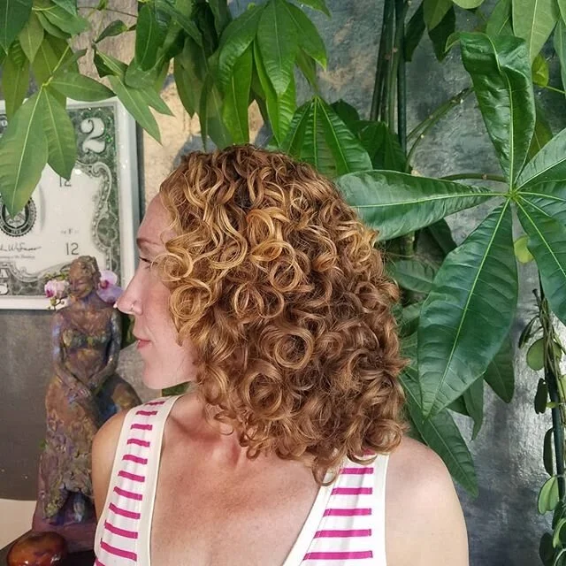A woman with curly, shoulder-length red hair, wearing a white tank top with pink stripes, standing in front of green leafy plants and a framed dollar bill.