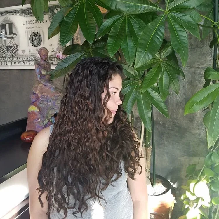 A woman with long, curly brown hair looking to her right, standing among green indoor plants.
