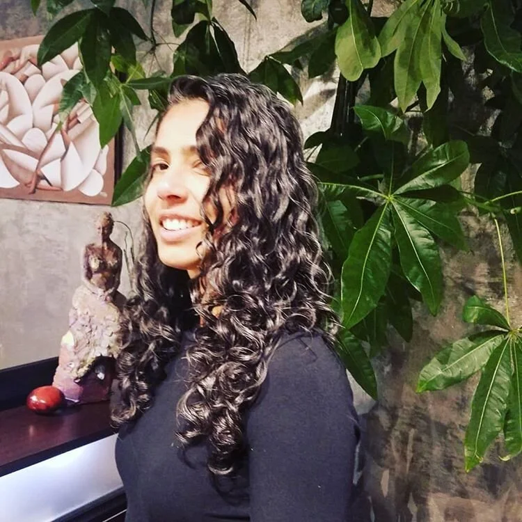 A young woman with long, curly dark hair smiling at the camera, standing indoors next to a large leafy plant, with a decorative sculpture and painting in the background.