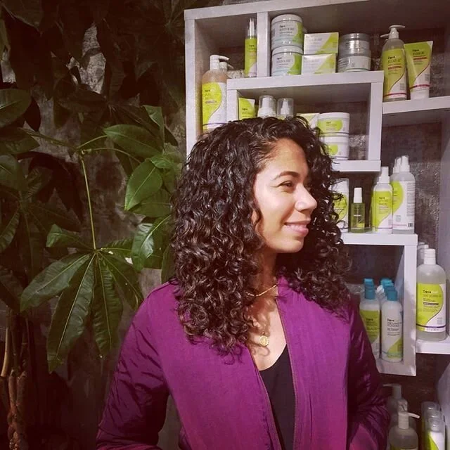 Woman with curly hair wearing a purple jacket standing in front of a shelf with skincare or haircare products.