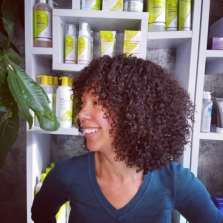 A woman with curly hair smiling, sitting in front of shelves filled with hair care products.