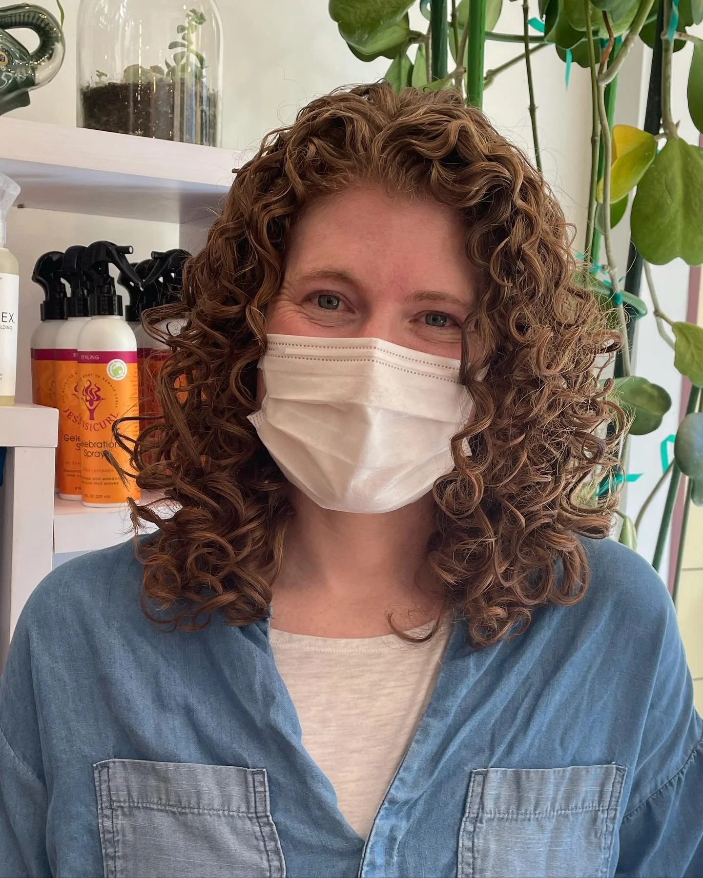 A woman with curly hair wearing a face mask and a denim shirt, sitting in a room with plants and hair care products in the background.