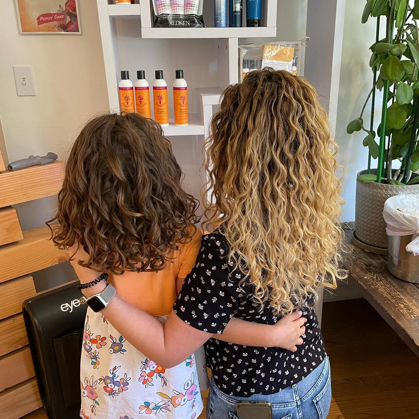 Two girls with curly hair embrace each other in a salon or store setting, one with darker brown hair and the other with blonde hair. Behind them are shelves with hair products and a large green plant.