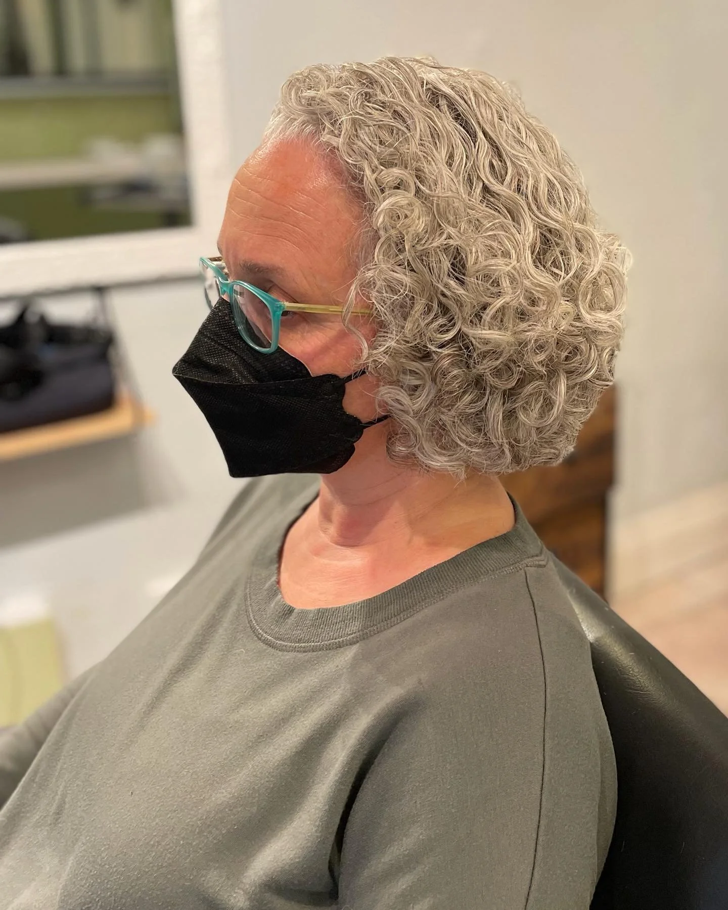 A woman with short, curly gray hair wearing glasses and a black face mask sitting in a chair.
