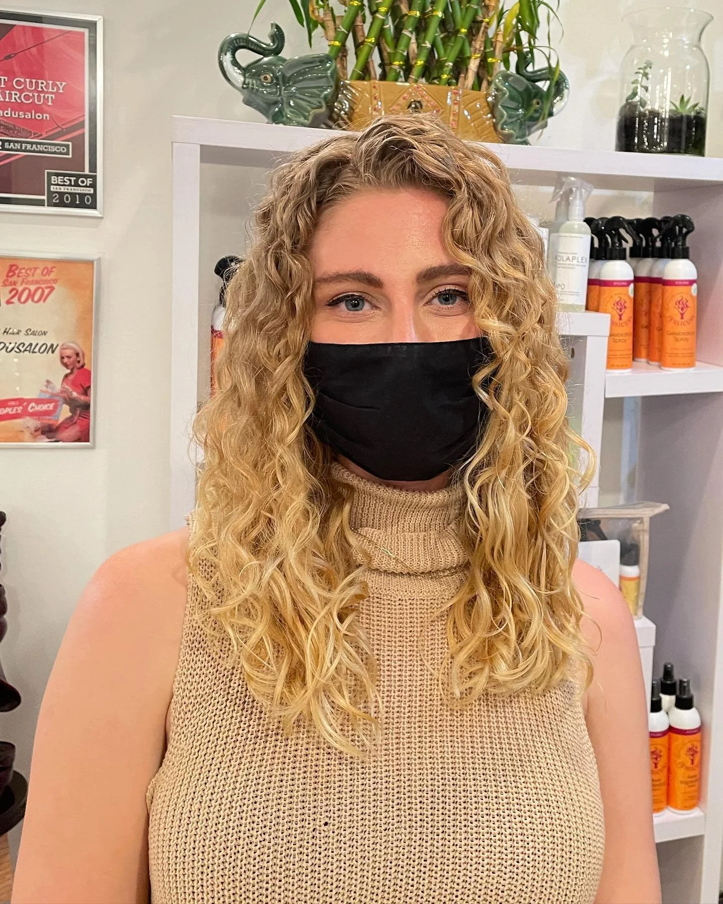 A woman with curly blonde hair wearing a black face mask and a tan sleeveless turtleneck sweater, standing inside a salon with hair products on shelves behind her.