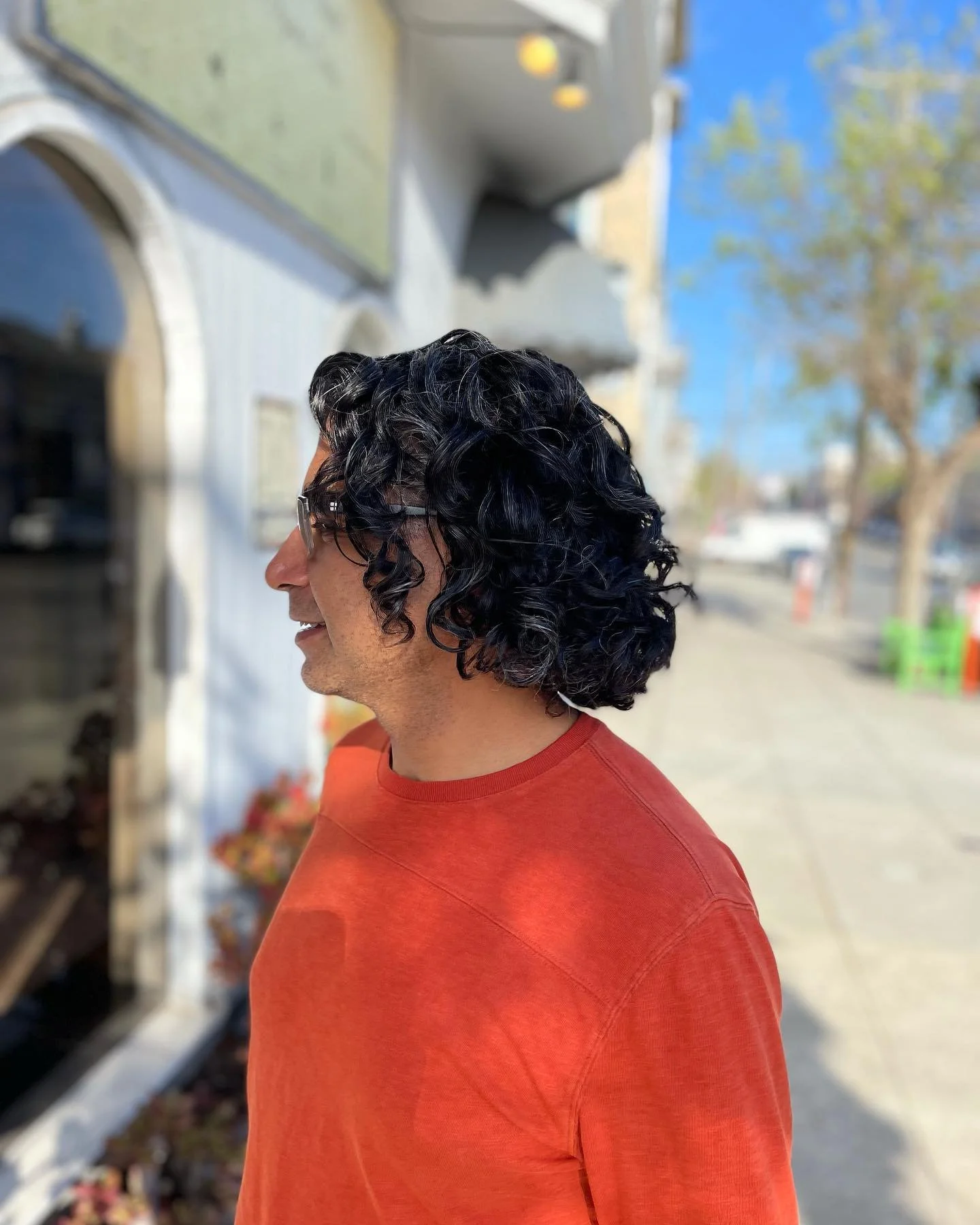 Side profile of a man with curly black hair, wearing sunglasses and a red shirt, standing on a sidewalk near a building with a blue sky in the background.