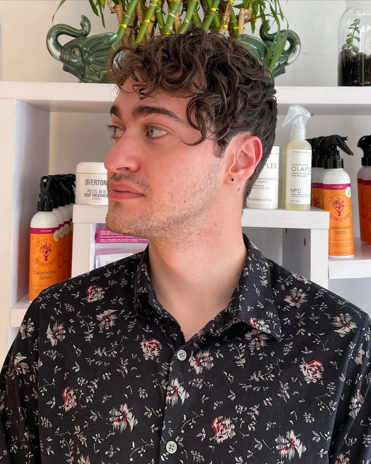 A young man with dark curly hair, wearing a black floral button-up shirt, standing indoors against a background of shelves with hair care products and a potted bamboo plant.