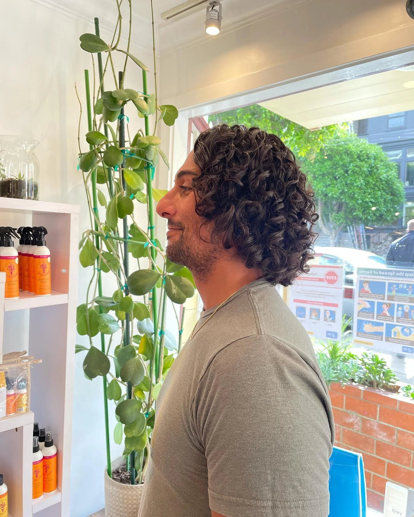 A man with curly dark hair and a beard standing inside a store, near a tall potted plant with broad green leaves, next to shelves with orange spray bottles and other products, with a window behind showing greenery and signage outside.