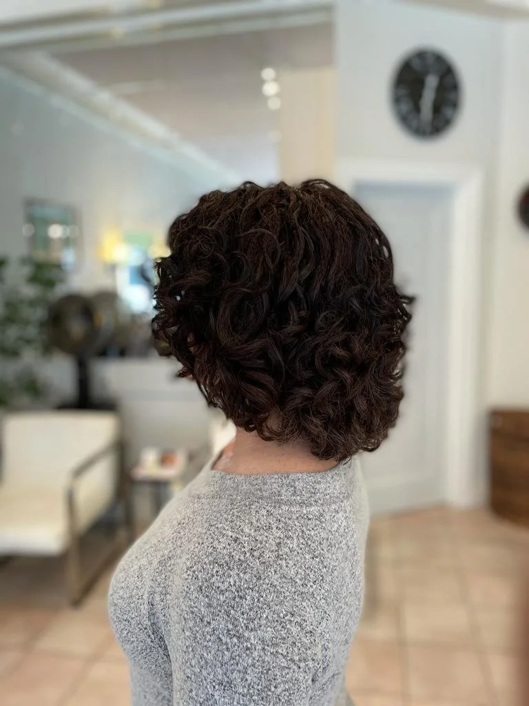 Back view of a person with short, dark, curly hair indoors, wearing a light-colored sweater.