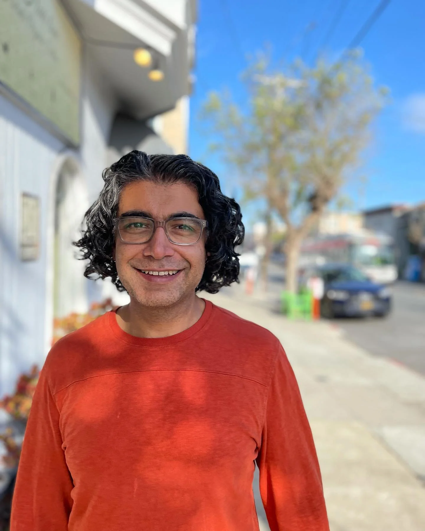 A man with black curly hair, glasses, and a bright smile, wearing an orange shirt, standing outside on a sunny day with a street, some trees, and buildings in the background.