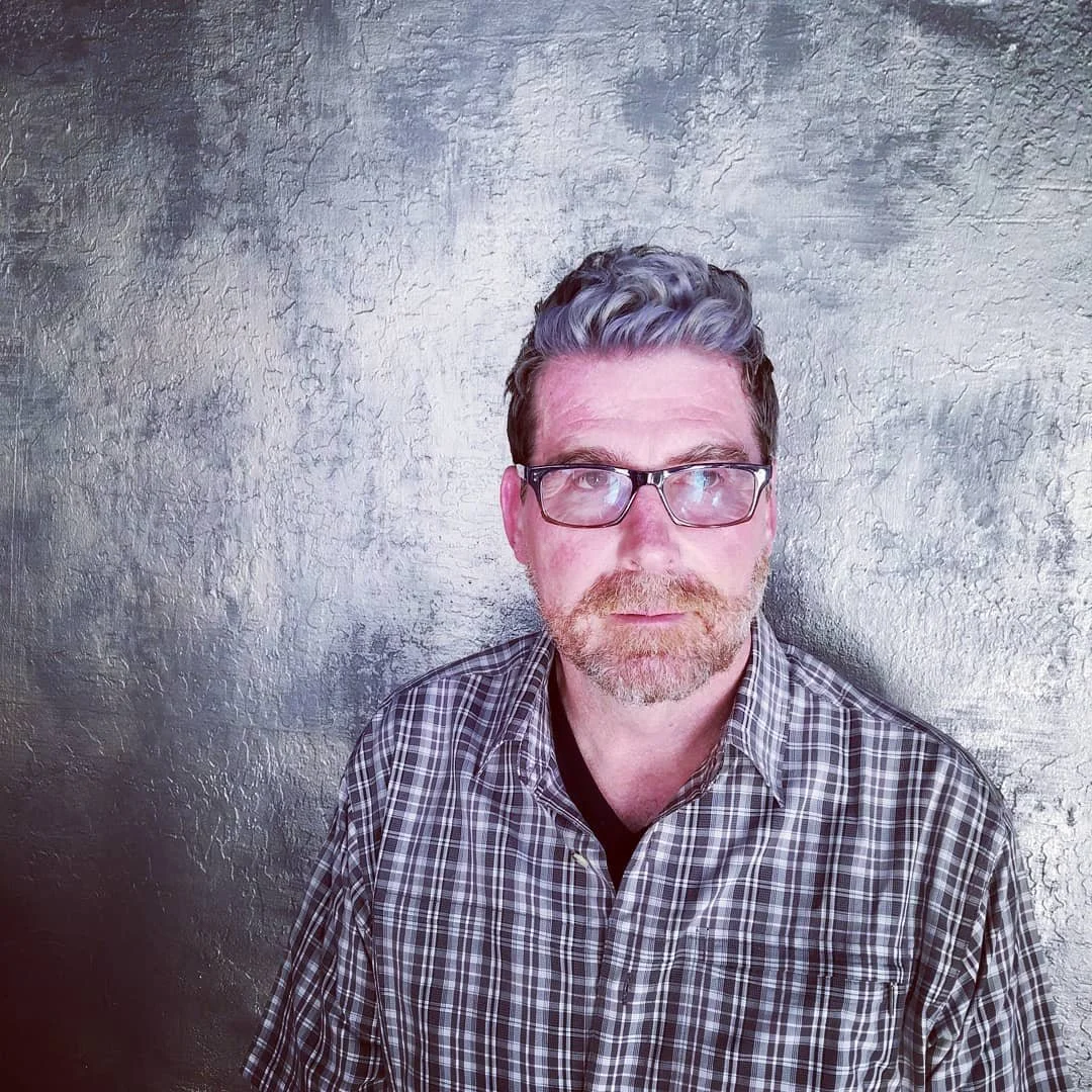 A man with glasses and a beard wearing a checked shirt standing in front of a textured metallic wall.