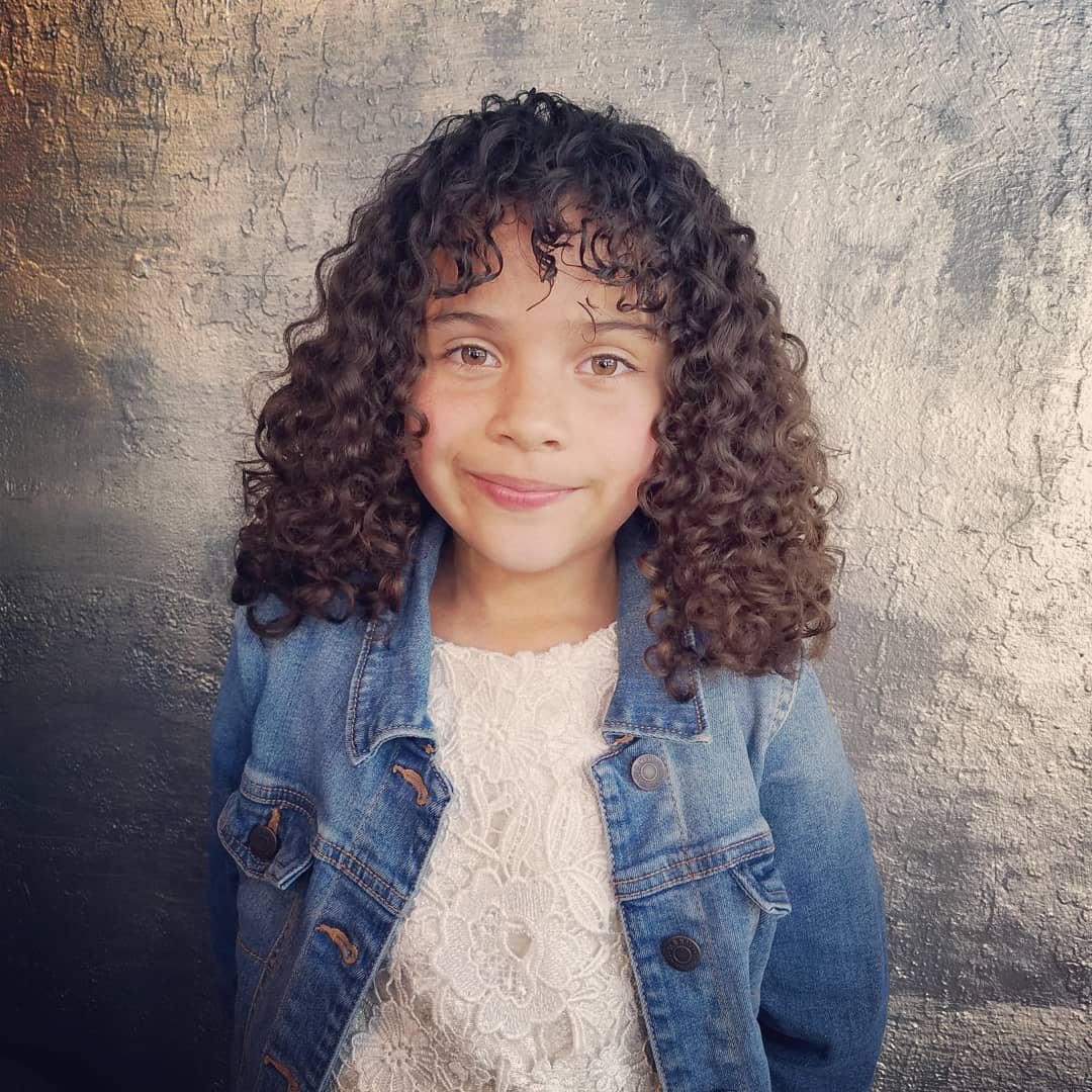 A young girl with curly brown hair, smiling, wearing a white lace top and denim jacket, standing against a textured gray wall.