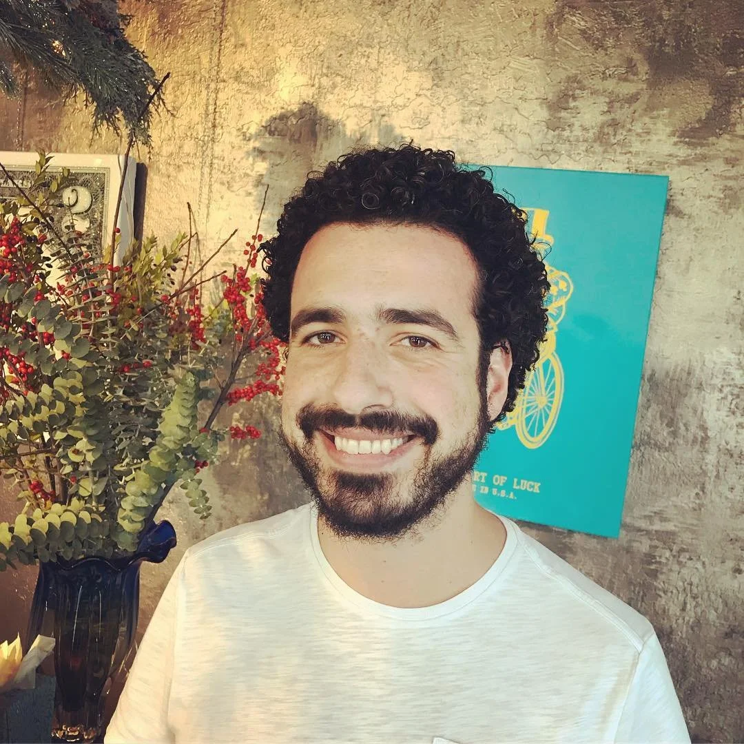 A smiling man with curly black hair, a beard, and mustache, wearing a white t-shirt, standing indoors near a floral arrangement with red berries and green leaves, and a blue poster with a yellow bicycle illustration in the background.