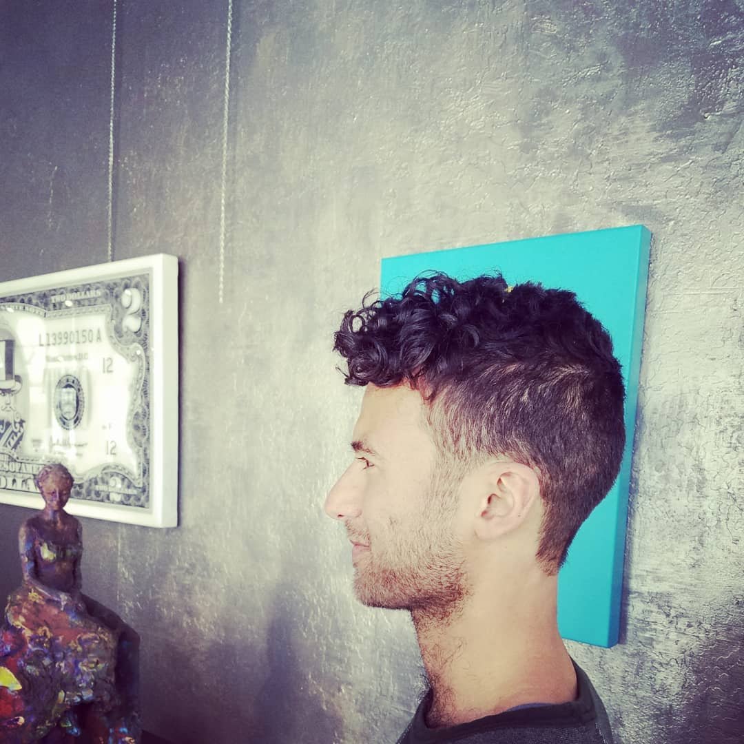Profile of a young man with curly hair and a beard, standing against a wall with artwork and a dollar bill framed. There is a colorful sculpture of a seated woman in front of him.