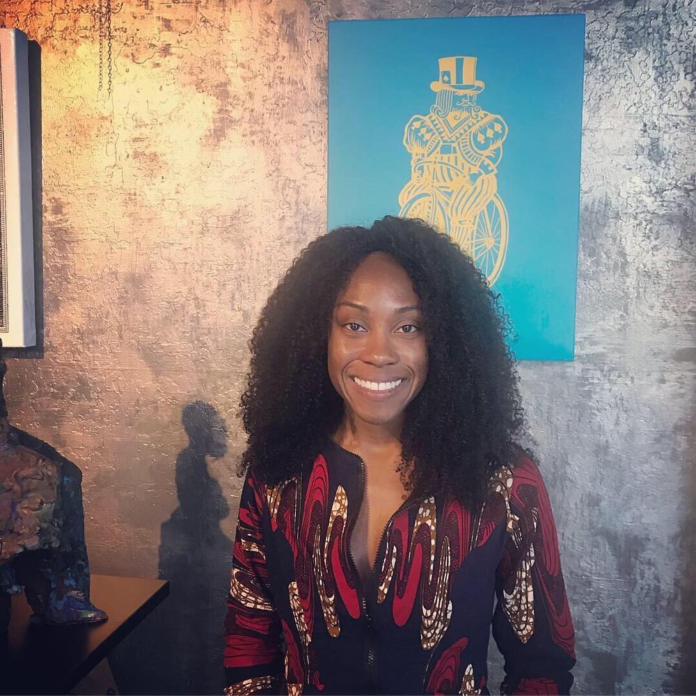 A smiling woman with curly black hair standing in front of a textured gray wall with a blue artwork featuring a cyclist wearing a top hat.
