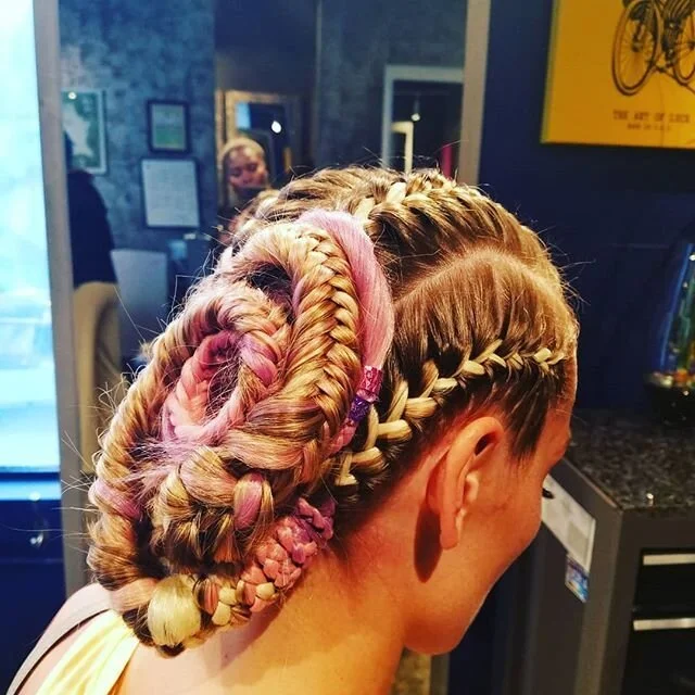 Side view of a woman with an intricate hairstyle consisting of braided and twisted hair, with some sections highlighted in pink and blonde, in a kitchen or salon setting.