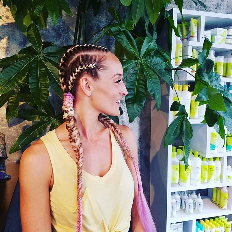 A woman with braided hair and pink tips, wearing a yellow tank top, smiling and looking to the right, standing in front of green leafy plants and a shelf filled with bottles.
